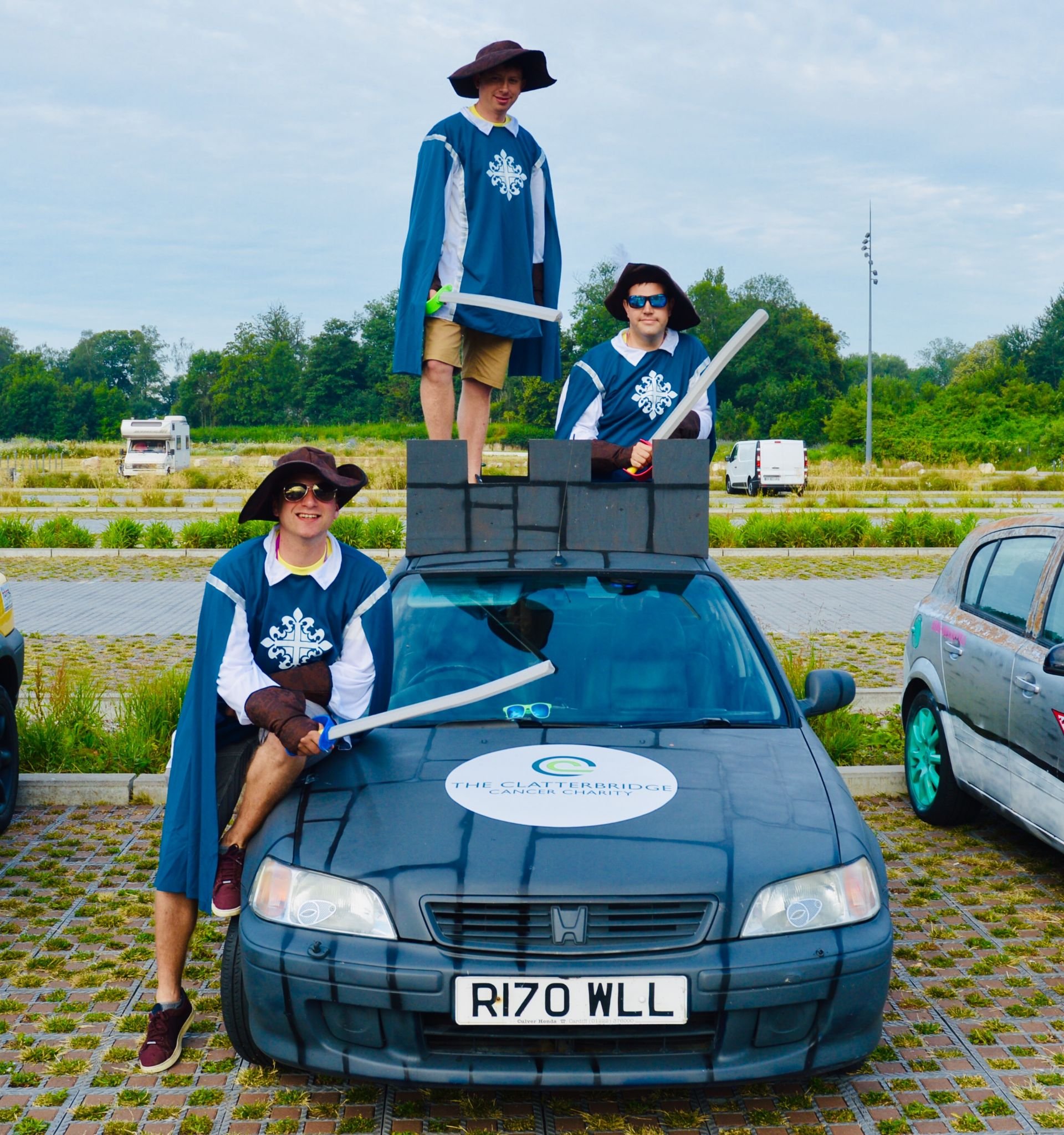 Three people dressed as fantasy knights with swords, standing on and around a car in an outdoor parking lot, with a sign for Cancer Charity on the car, and a rural landscape in the background.