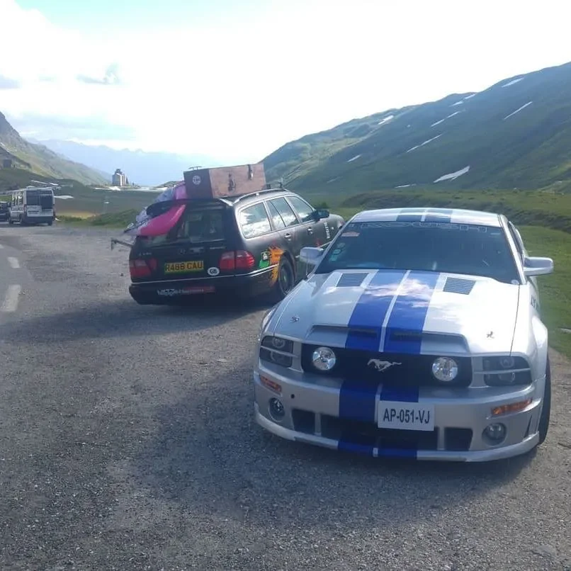 A roadside scene with two parked cars on a mountain road, surrounded by green hills and snow patches, with other vehicles in the background.