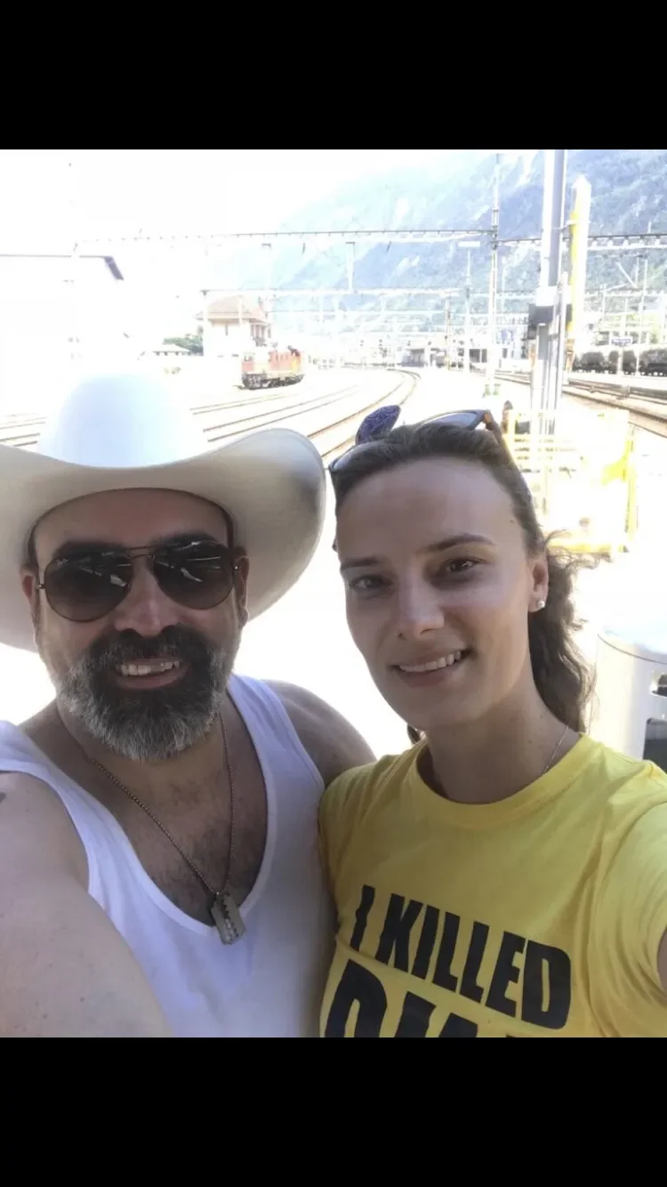 A man and woman taking a selfie at a train station. The man is wearing sunglasses, a large white cowboy hat, a white tank top, and a necklace. The woman is wearing a yellow T-shirt with black text and has sunglasses on her head. Background shows trai