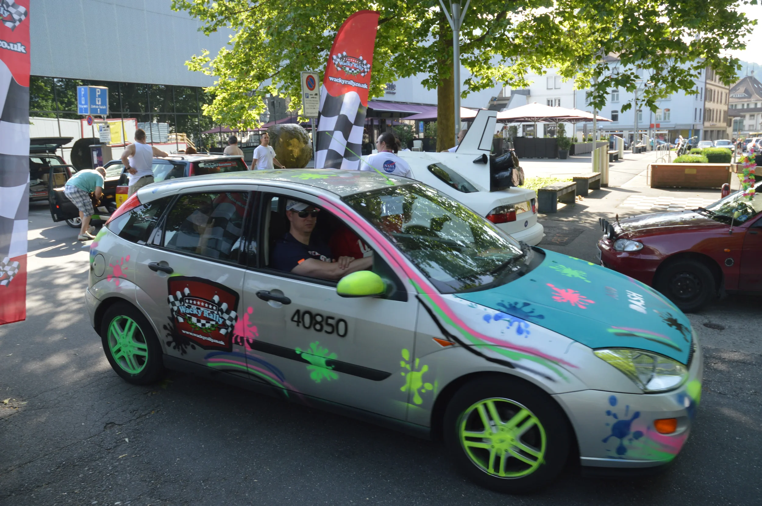 A silver hatchback car decorated with colorful paint splashes and Wacky Rally decals, parked on a city street with other cars and people around.