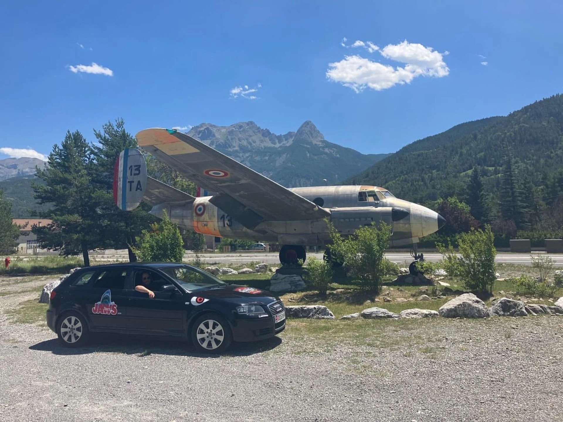 A vintage fighter jet airplane displayed outdoors behind a black car with a 'Jaws' sticker, with mountains and trees in the background under a blue sky with some clouds.