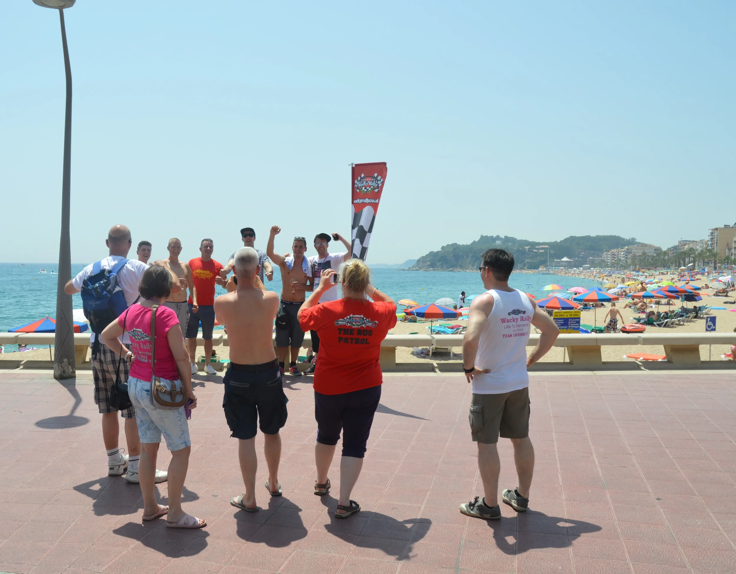 A group of people stands on a beach promenade facing a panel of speakers, with a busy beach full of umbrellas and sunbathers in the background. The sky is clear and blue.