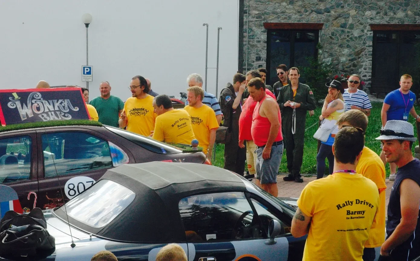 A group of people gathered around rally cars, with some wearing yellow t-shirts that say 'Rally Driver Barmy to Barcelona,' at an outdoor event.