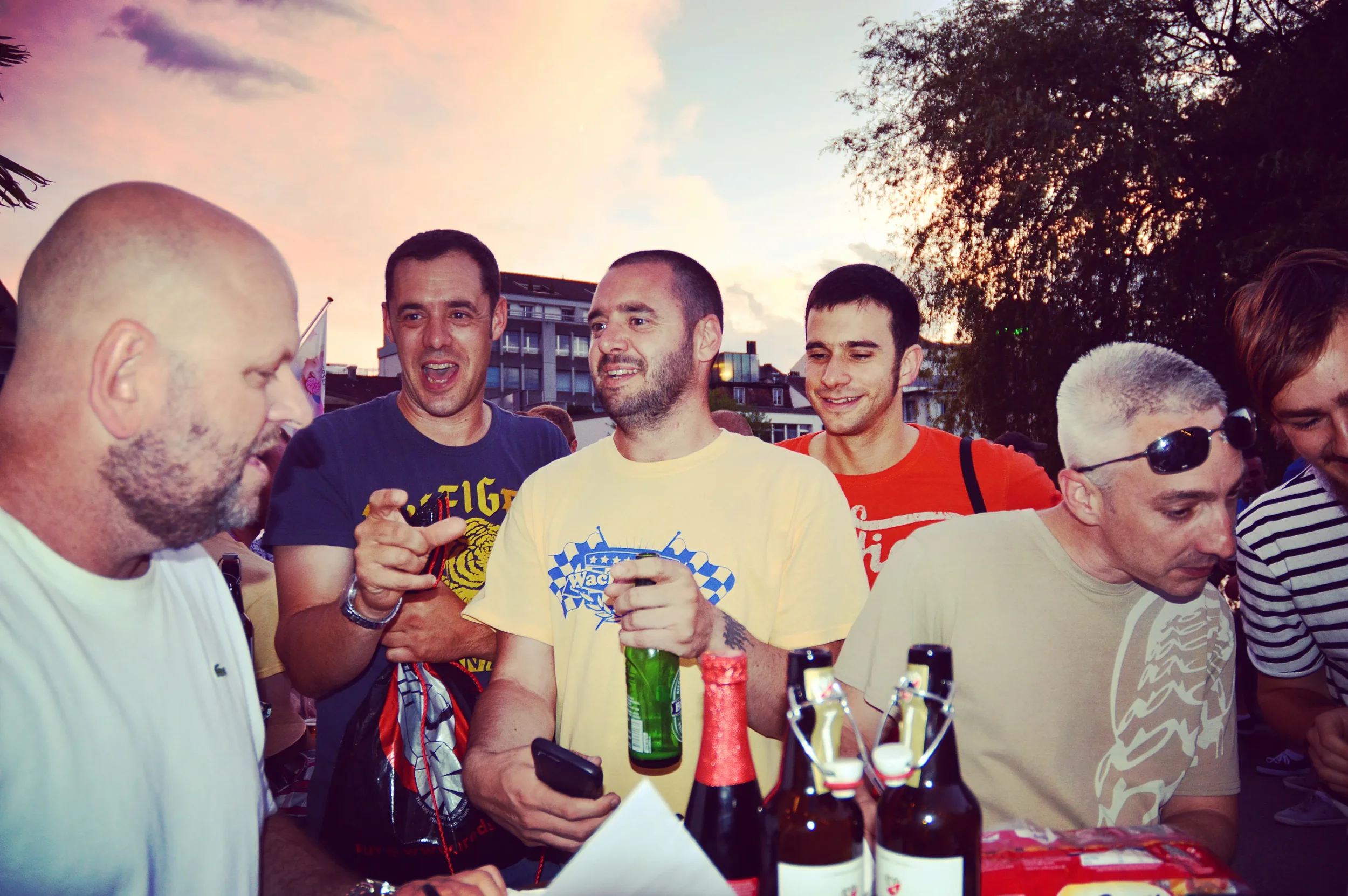 A group of men gathered outdoors at sunset, socializing with drinks and bottles on a table, some smiling and engaged in conversation.
