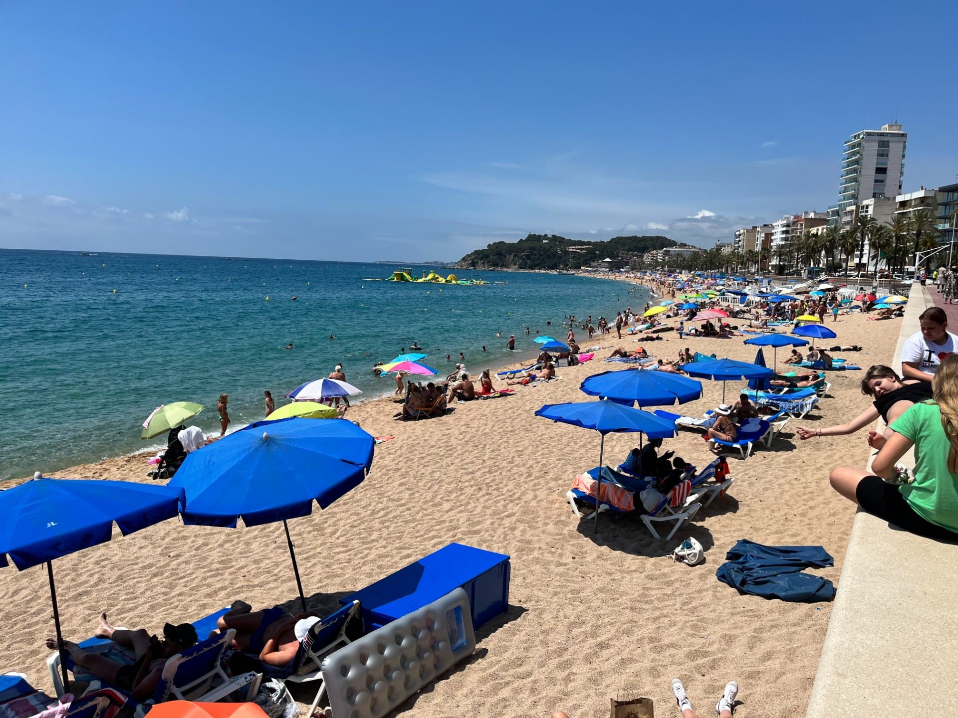 Crowded beach with colorful umbrellas, people swimming, sunbathing, and walking along the shoreline. Tall buildings and palm trees line the background under a clear blue sky.