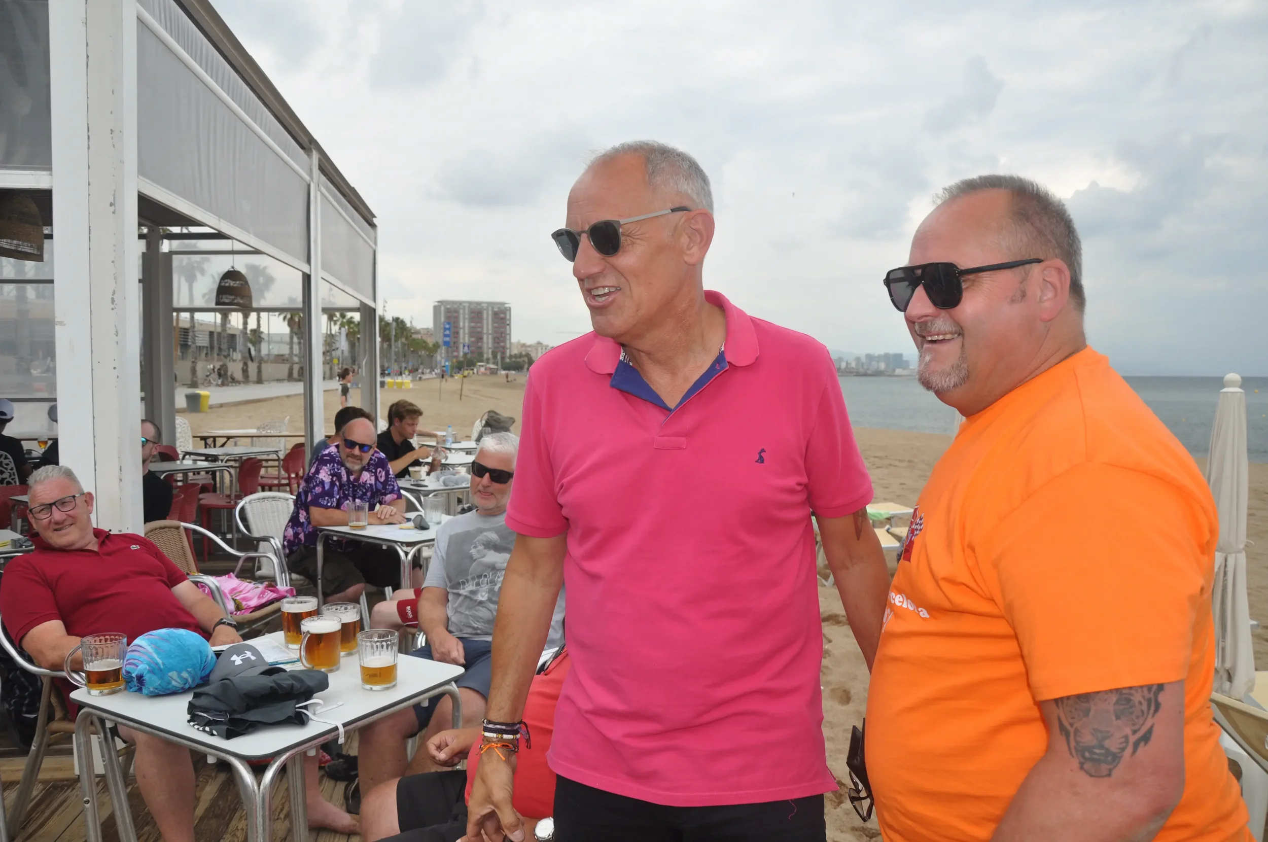Group of men enjoying drinks at a beachside restaurant, some wearing sunglasses, with others sitting at tables behind them on the sand, and the ocean and cloudy sky in the background.
