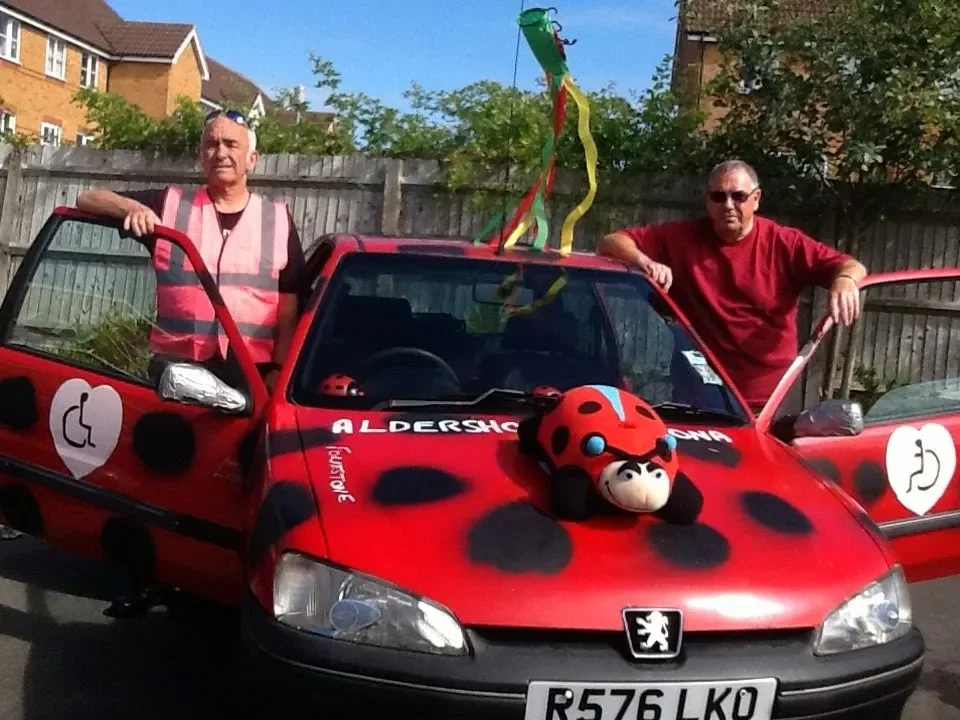 Two men standing by a red car decorated with ladybug-themed accessories and symbols, including a ladybug plush on the hood and streamers on top, in a residential outdoor setting.