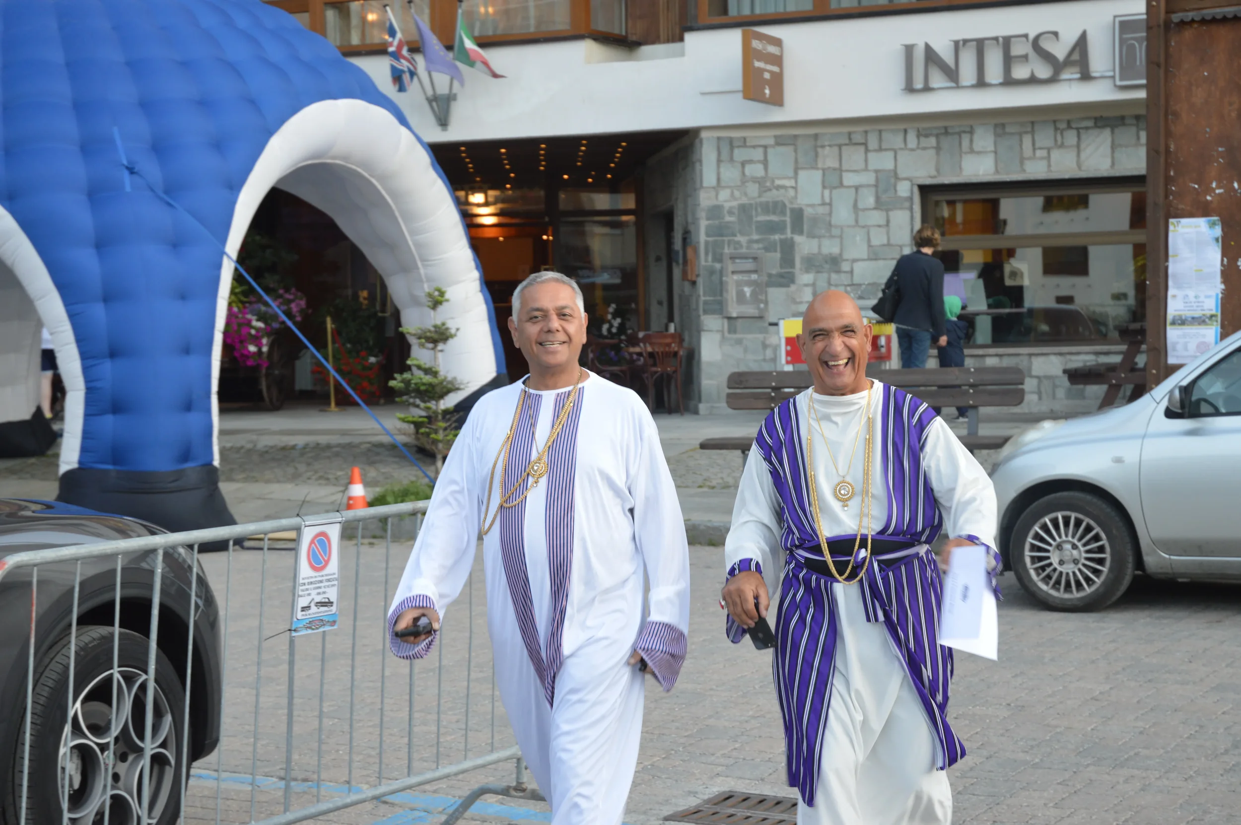Two men in traditional Ethiopian clothing, smiling and walking outdoors in front of a building with a sign that reads 'INTESA'. An inflatable arch and flags are visible in the background, with a few people and parked cars nearby.
