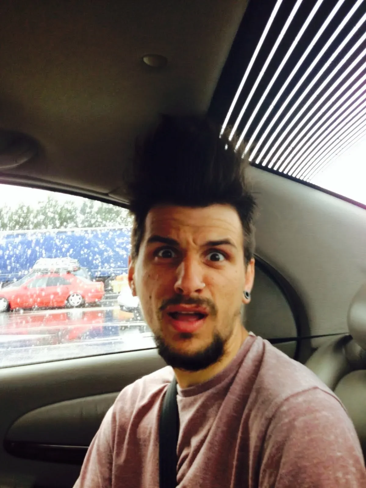 A man with dark hair and facial hair making a surprised expression while sitting in the backseat of a car, with red and blue cars visible through the window during rainy weather.