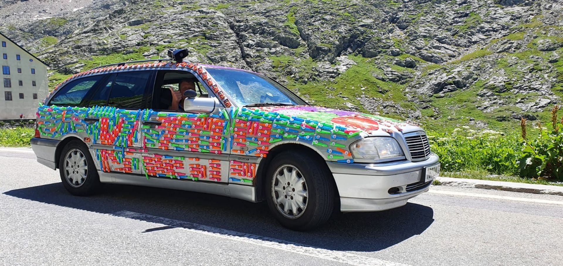 A car decorated with colorful plastic straws arranged to spell out 'ALWAYS' on the side, with additional designs on the hood, driving on a mountain road with green and rocky terrain in the background.