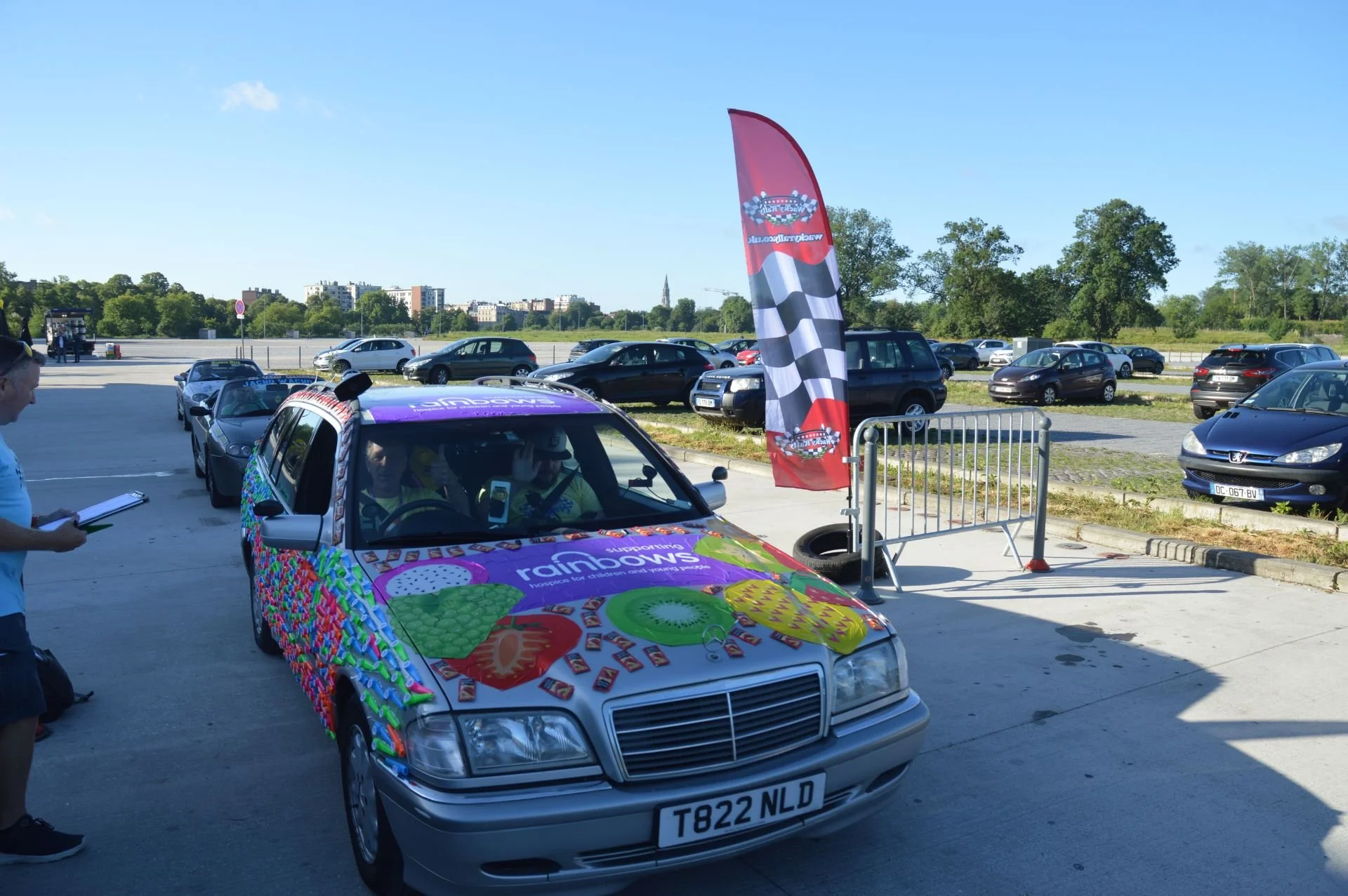 A decorated Mercedes-Benz car with colorful fruit and candy-themed decals parked in an outdoor lot next to a checkered flag banner, with a person holding a clipboard nearby, and several other cars parked in the background on a sunny day.