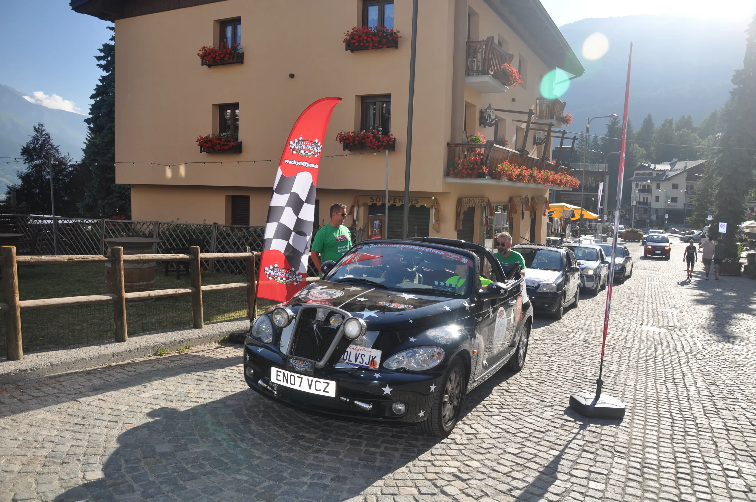 A small black car decorated with white stars, participating in a car rally with Italian license plates, is parked on a cobblestone street in a European town. There are people around, a red checkered and racing flag banner nearby, and a tan building w
