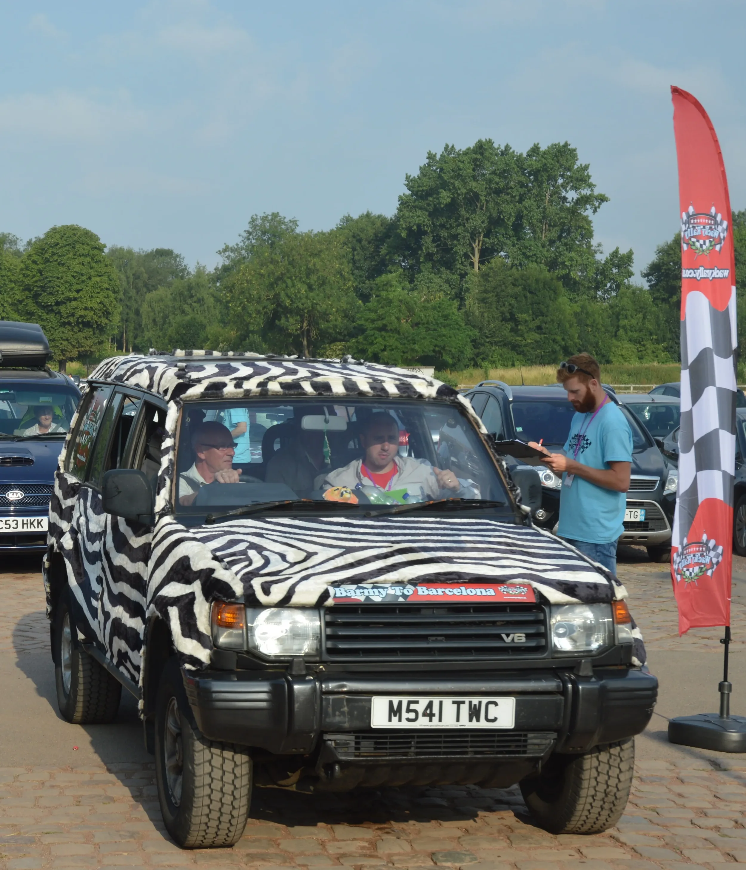 A black off-road vehicle with a zebra-patterned cover parked in a lot, with some people inside and someone outside writing on a clipboard. A red and black checkered flag with a logo stands nearby, and a banner on the vehicle reads "Barmy to Barcelona