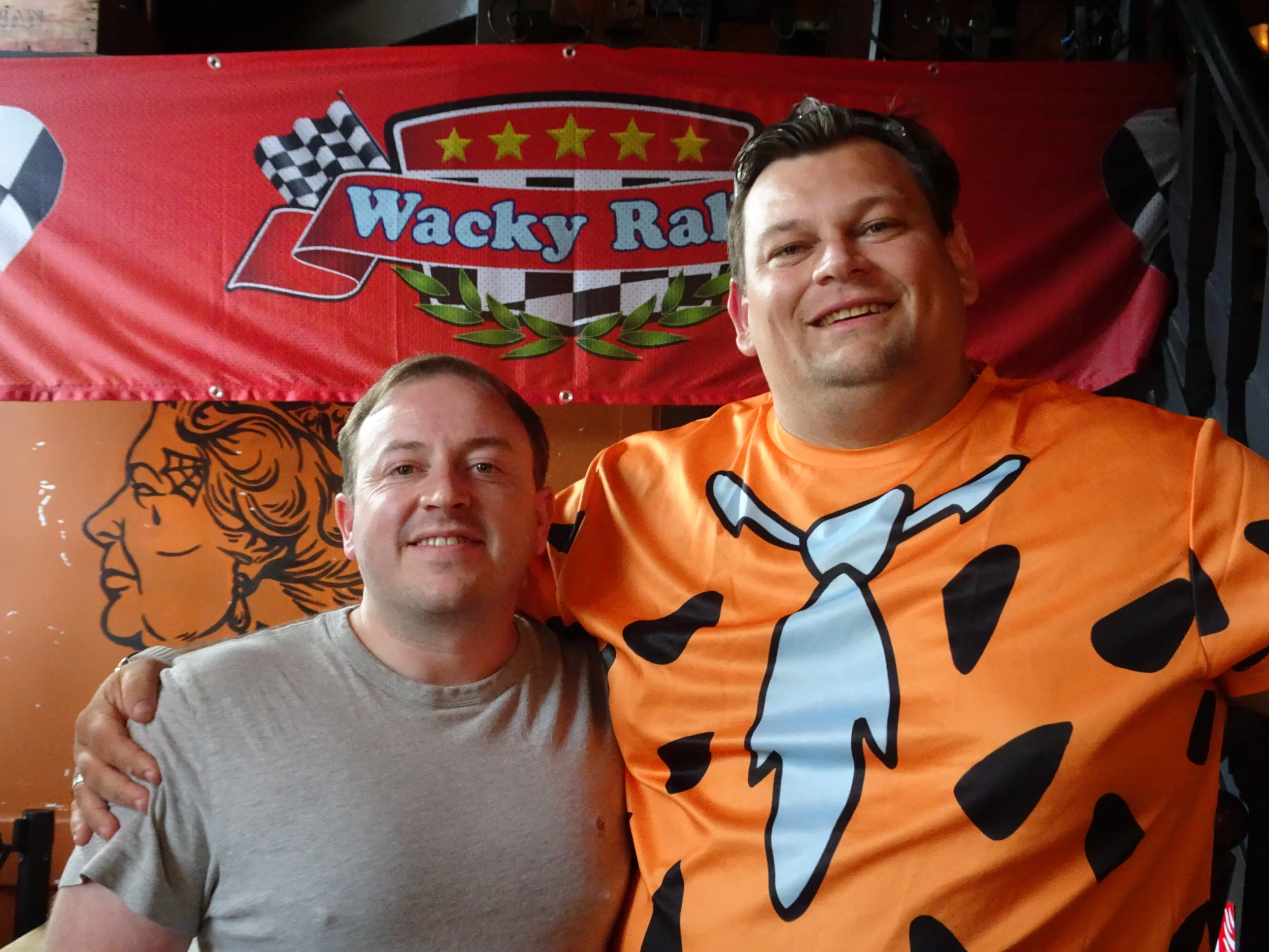 Two men smiling and posing together in front of a red banner with checkered flags and the words "Wacky Races". The man on the right wears a bright orange shirt with a cartoon-style cow print design.