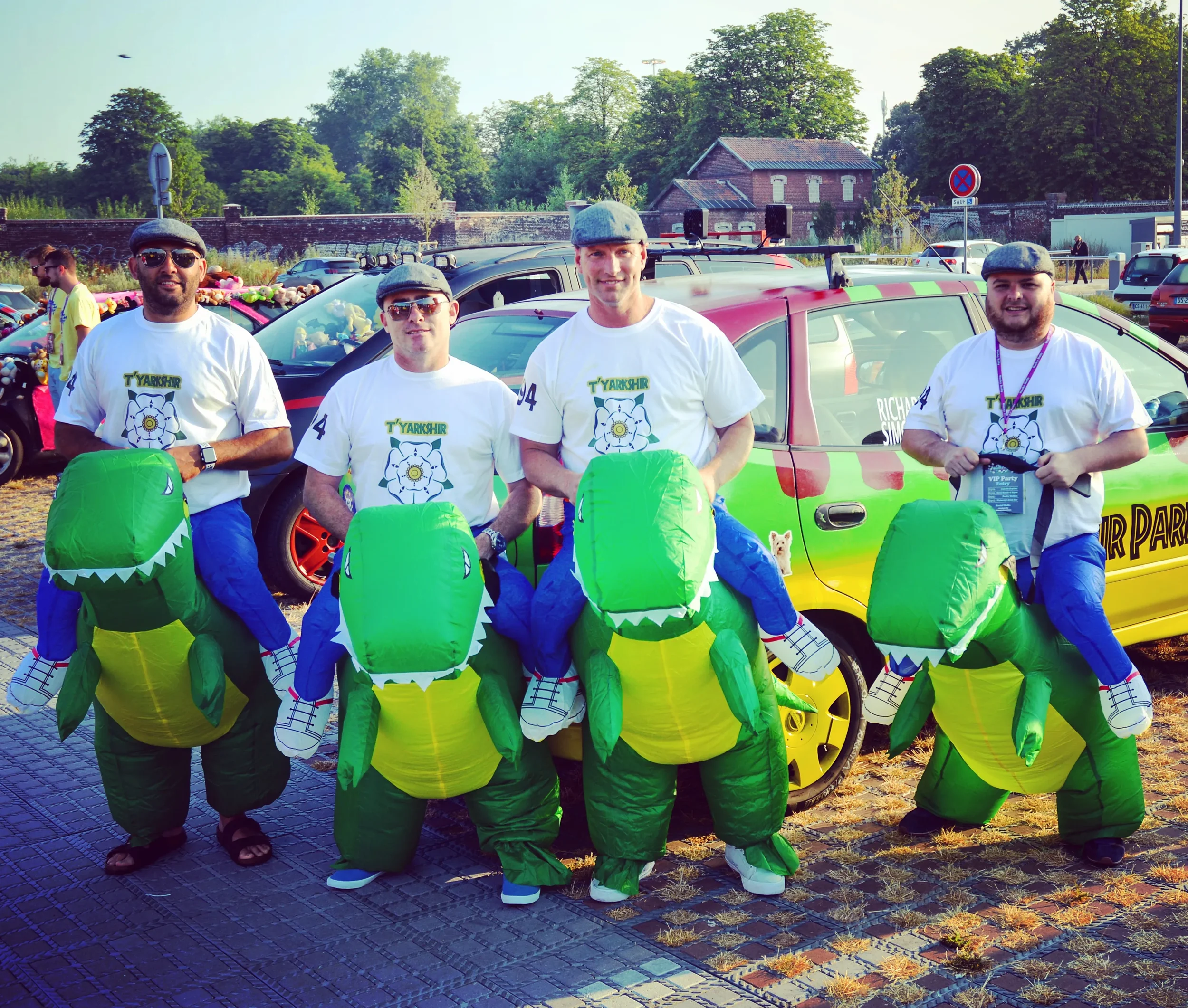 Four men dressed in T'YARKSHIR event shirts and blue pants wearing inflatable dinosaur costumes pose for a photo in a parking lot.