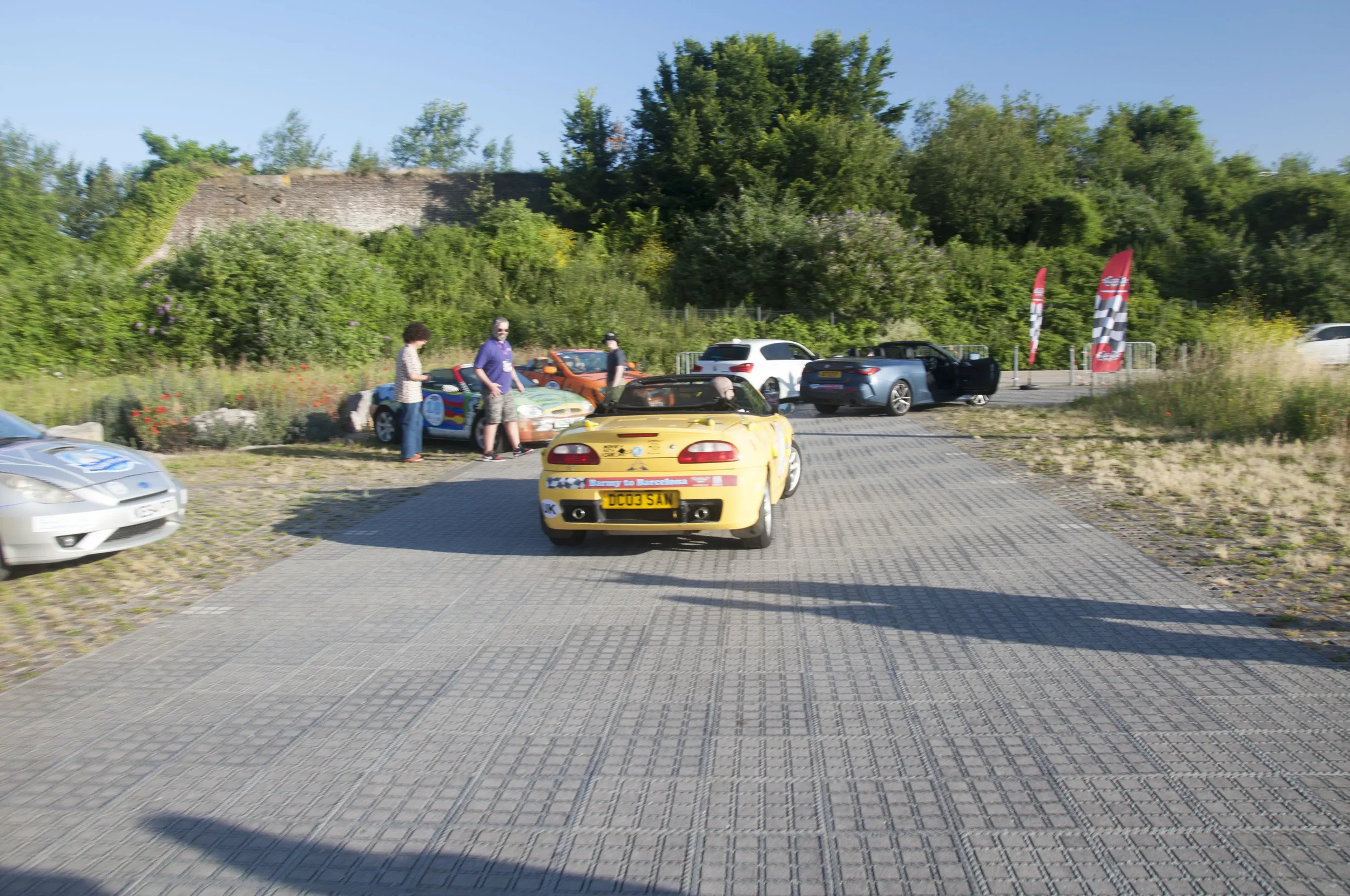 A parking lot with various cars, including a yellow convertible in the foreground and a group of people near some cars in the background, with trees and flags on the right side.