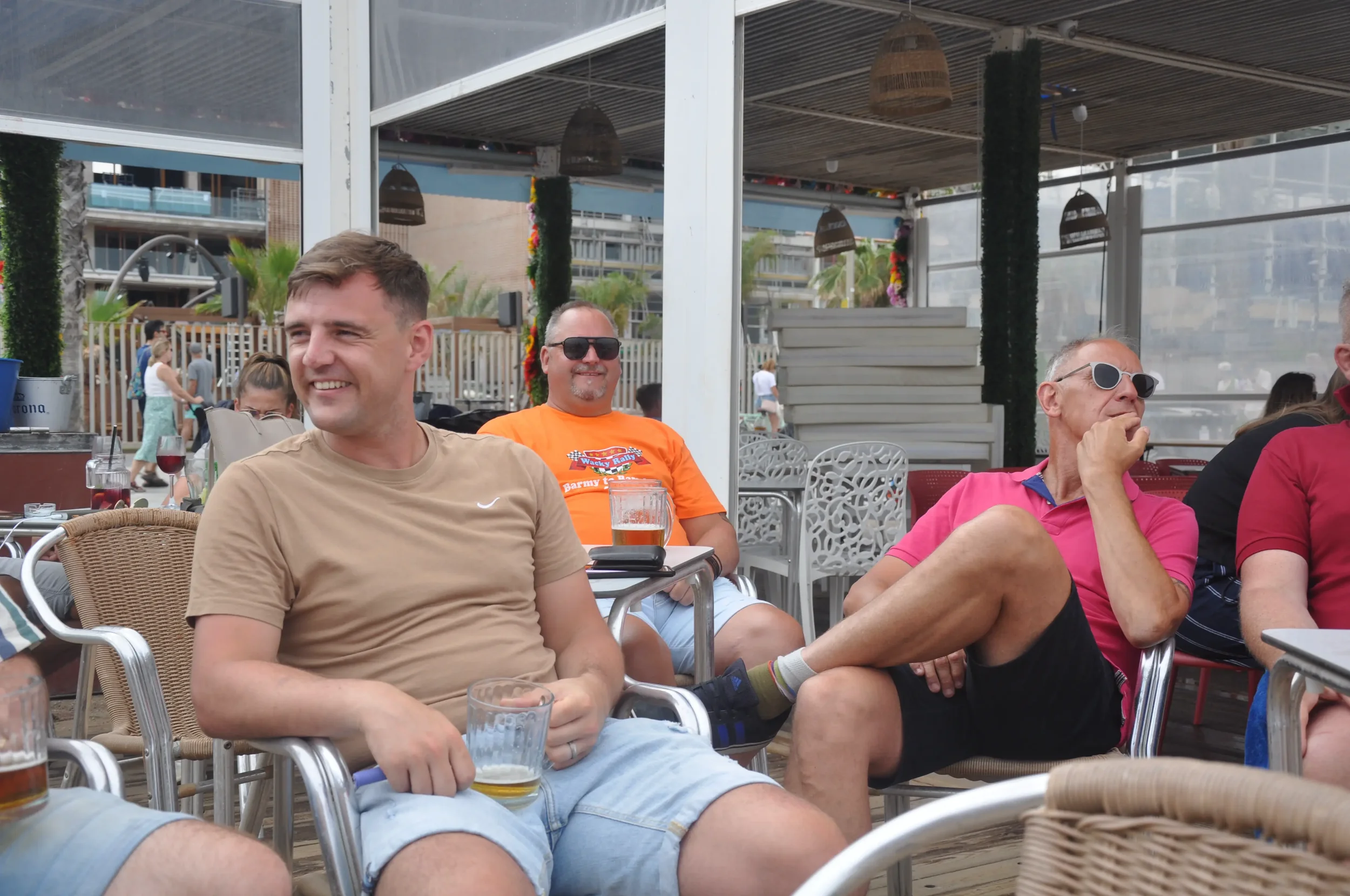 People sitting and relaxing at an outdoor venue, with drinks on tables, under a covered patio, in a lively, casual atmosphere.