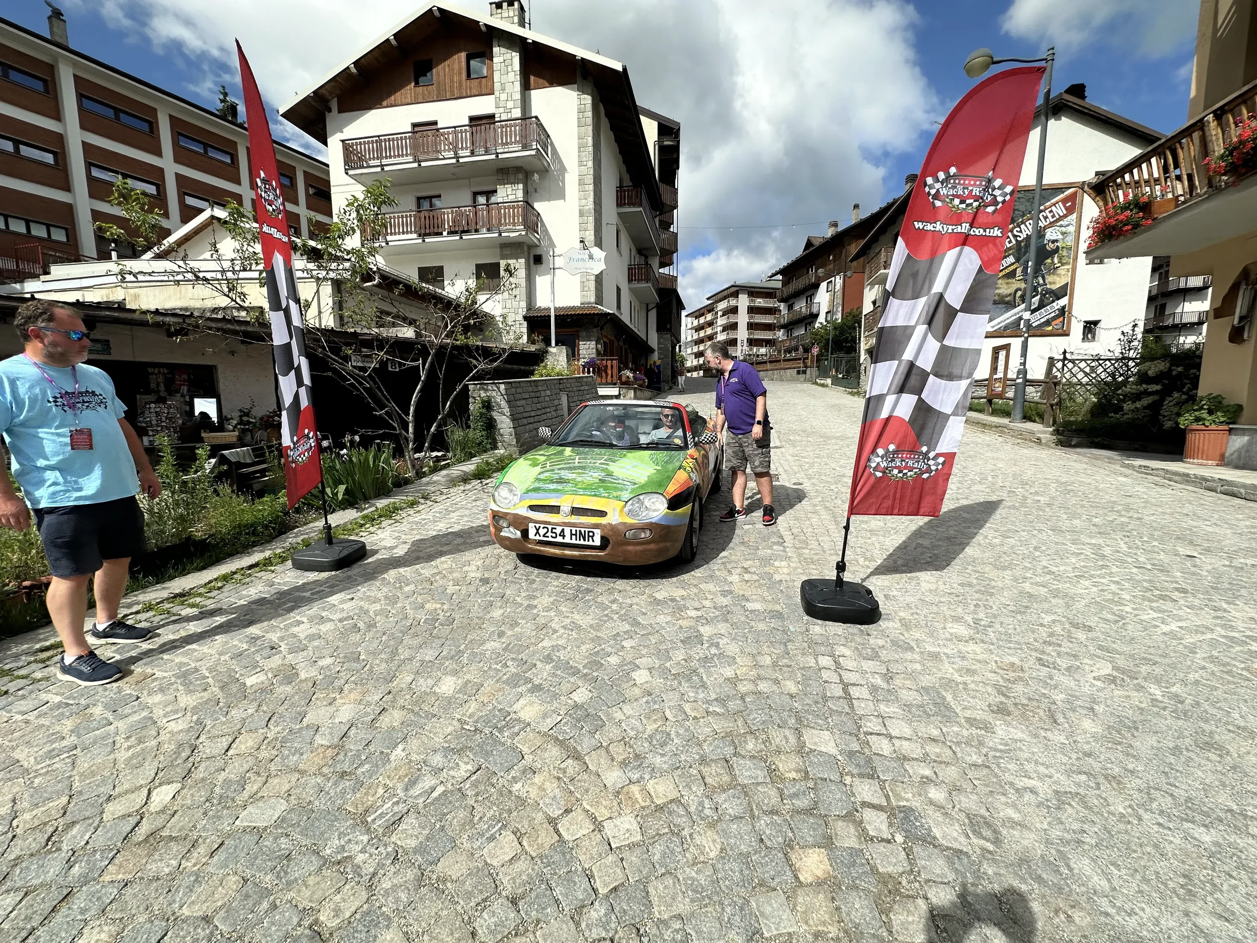 A small colorful car with racing flags on each side, parked on a cobblestone street in front of multi-story buildings. Two men are standing near the car, one looking inside, the other standing nearby. The scene is in a village or urban area with trad