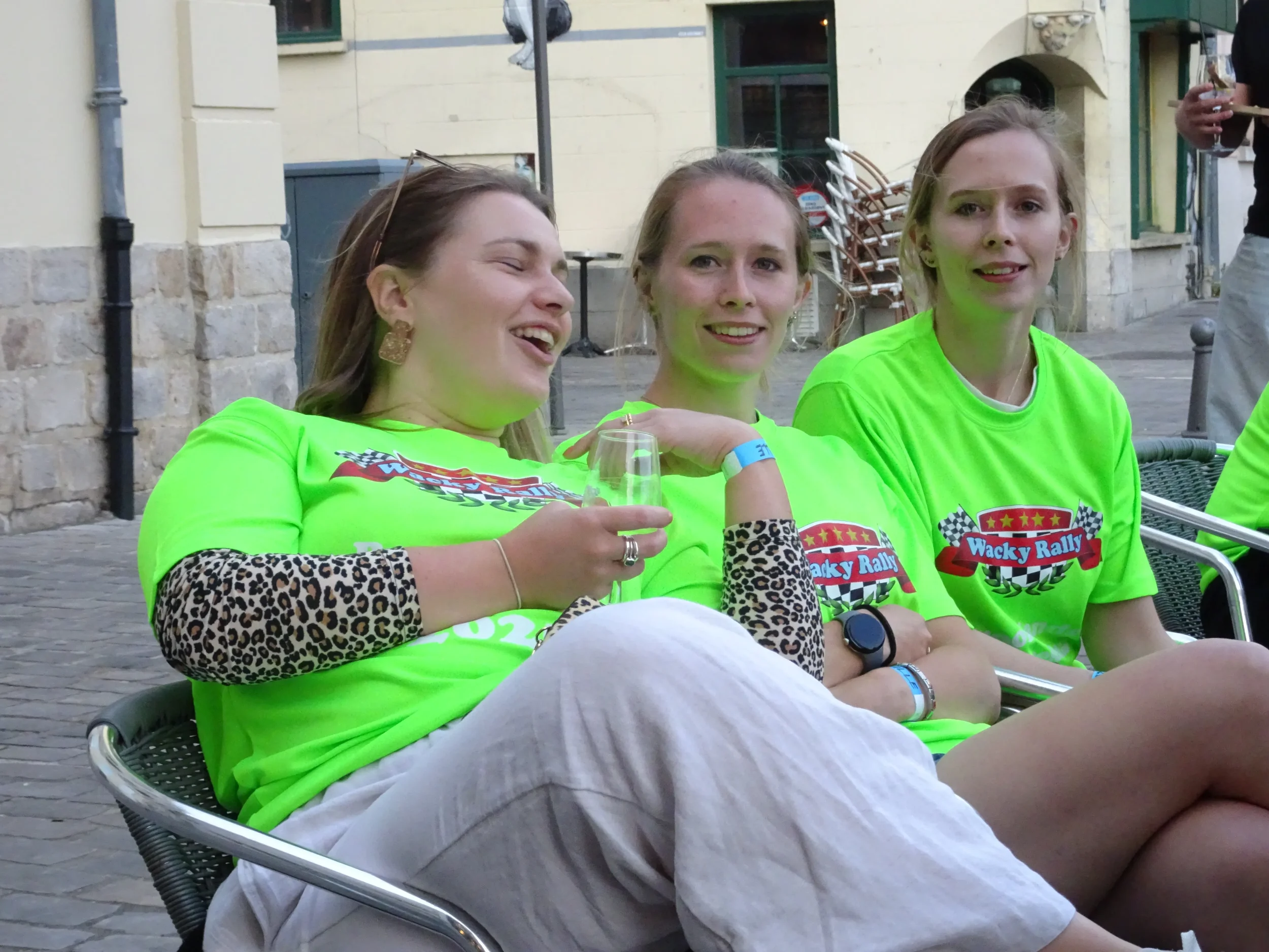 Three women sitting outdoors, wearing bright green T-shirts with a racing-themed logo, some smiling and one laughing, with one holding a glass of wine, at what appears to be a casual social gathering.
