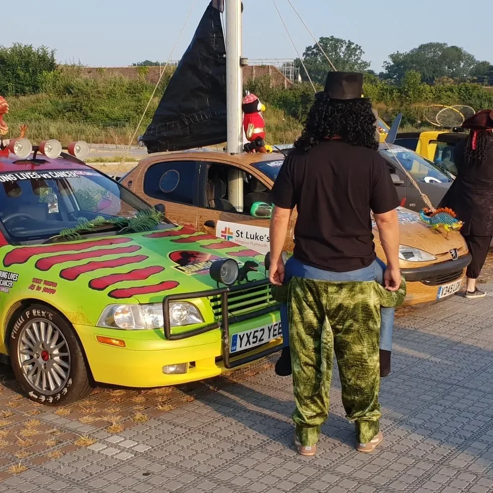 People dressed in costumes and face paint standing near decorated cars, possibly at a parade or festival.