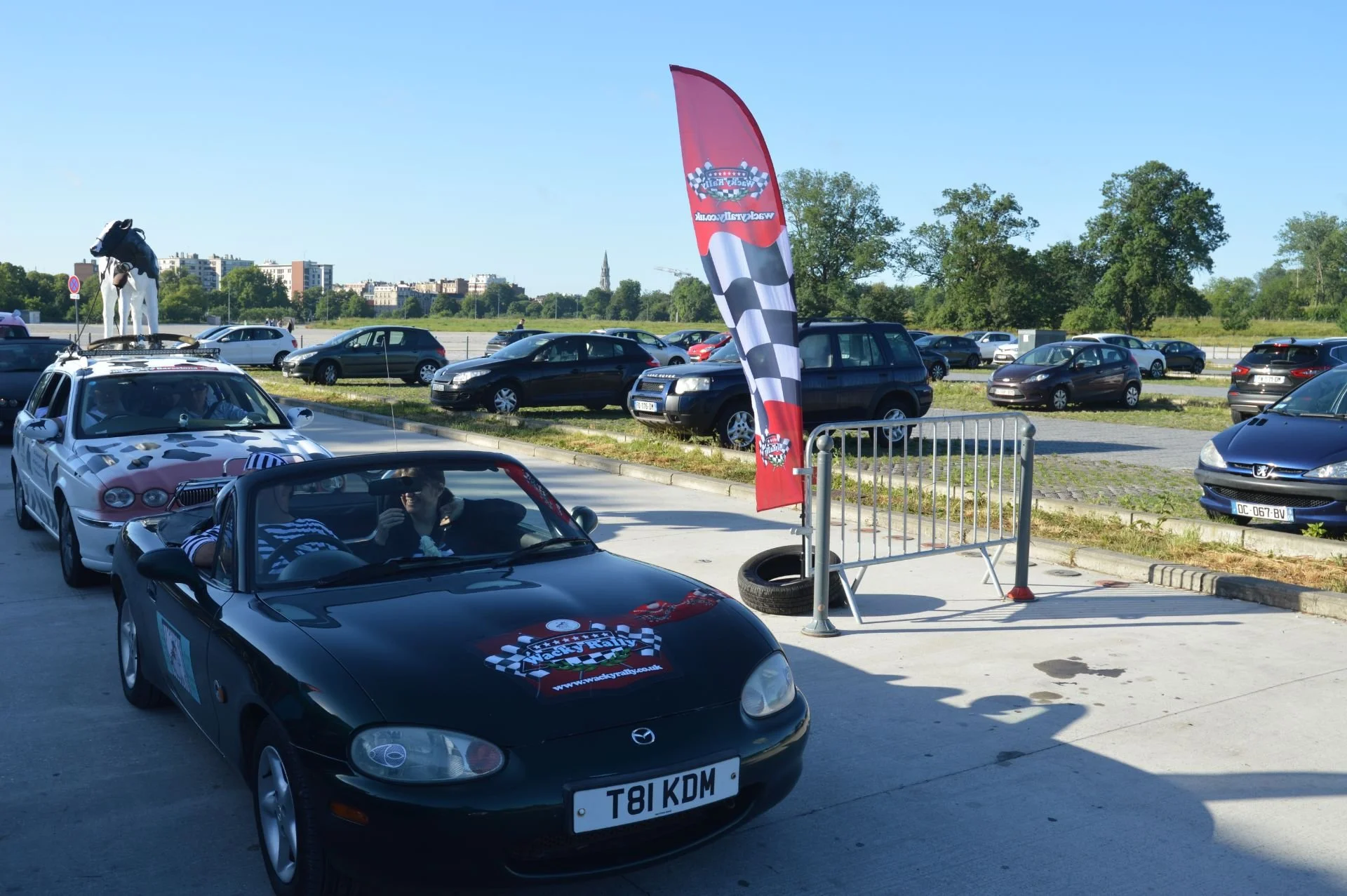 A black Mazda Miata convertible with a racetrack logo on the hood, parked outdoors on a sunny day, with people and cars in the background at a parking lot or event area.