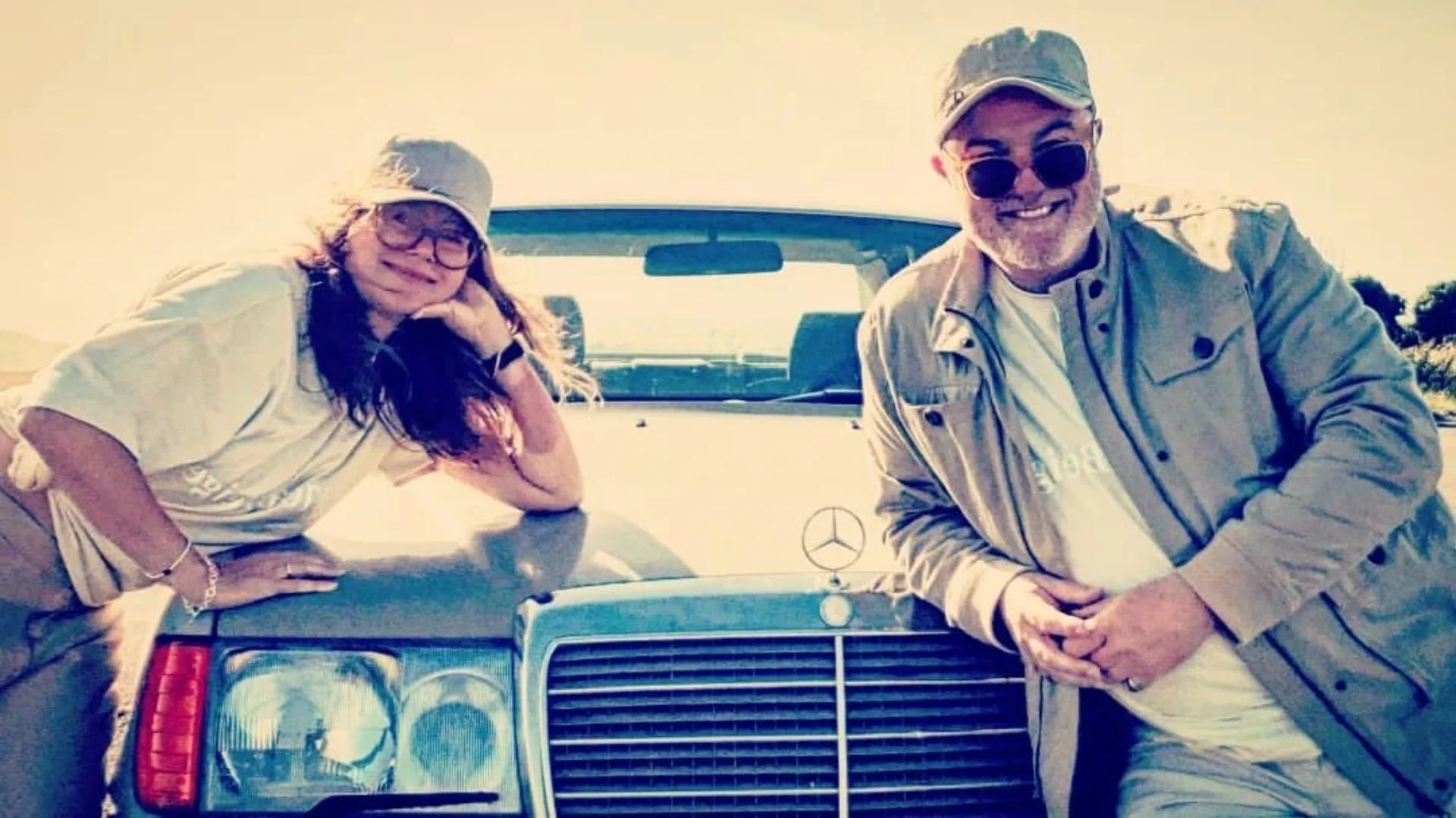 Two smiling adults, a woman and a man, leaning on a vintage silver Mercedes-Benz car parked outdoors on a sunny day.