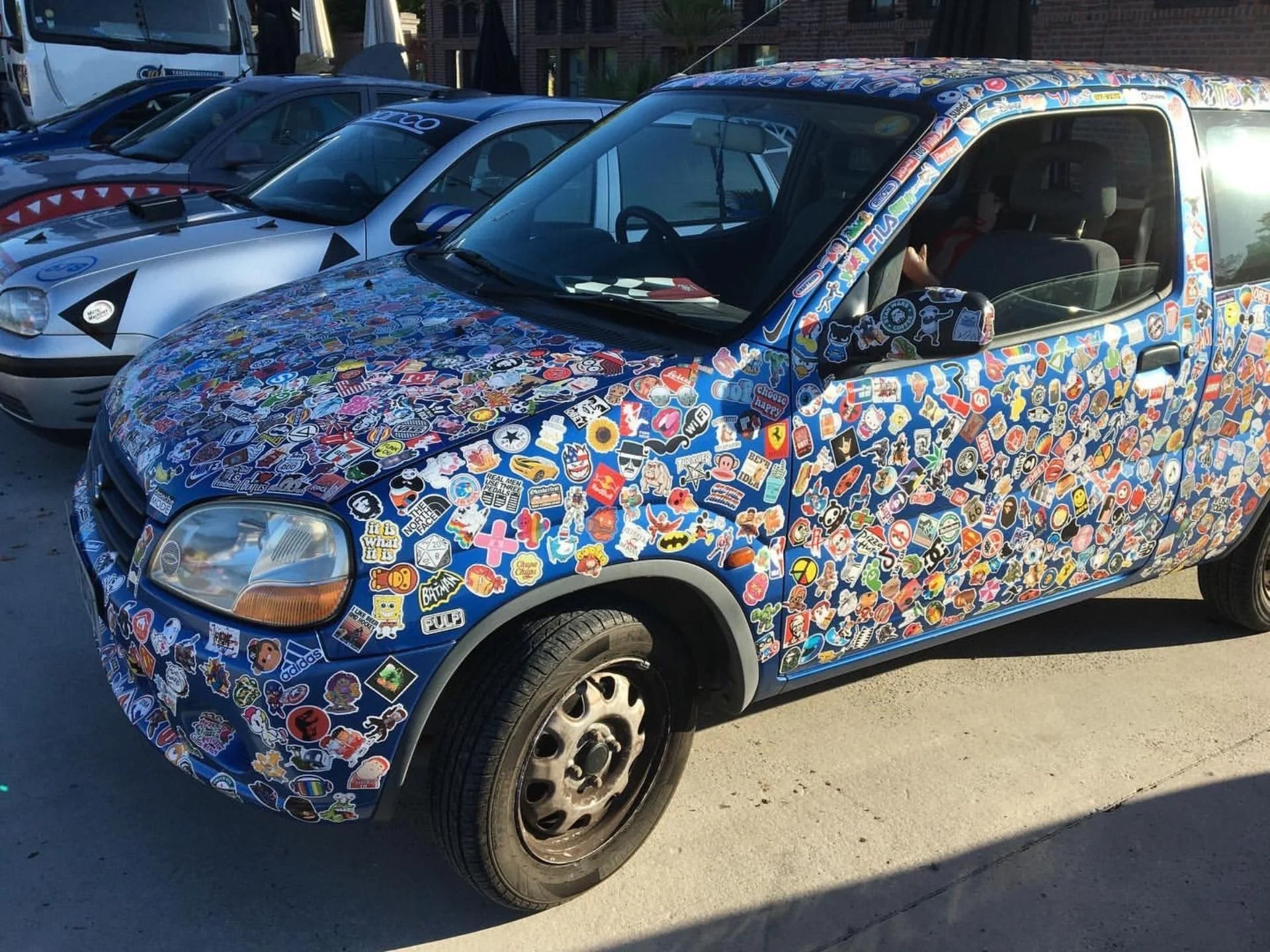 A blue car completely covered with various colorful stickers parked in a lot.