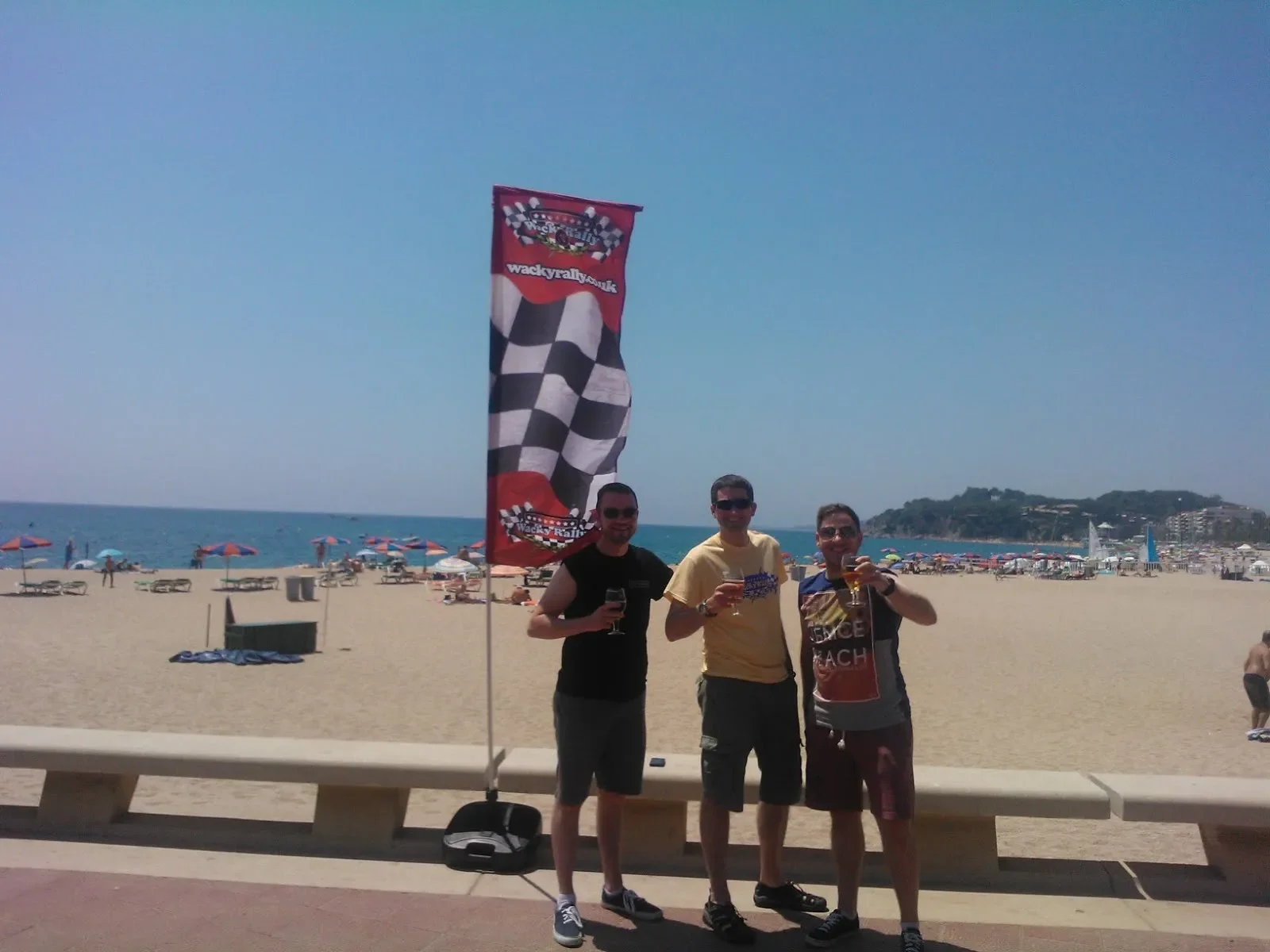 Three men holding drinks on a beach with colorful umbrellas, a flag with checkered design, and an ocean in the background.