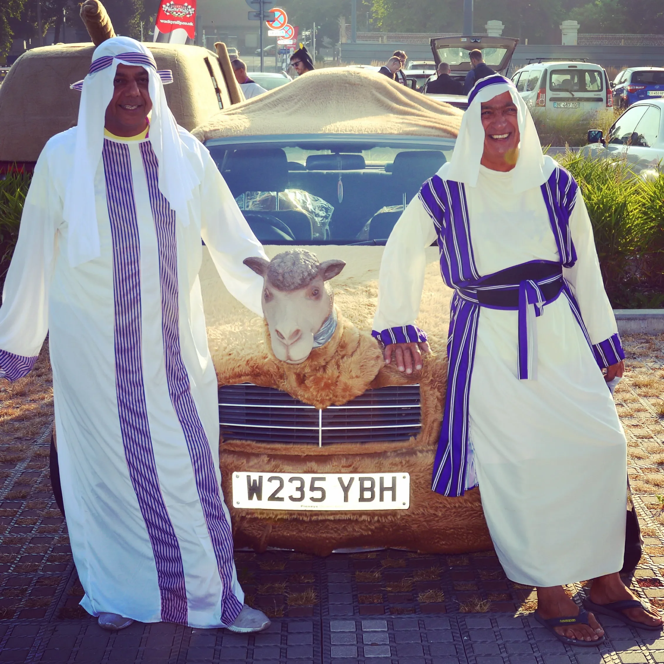 Two men dressed in traditional Middle Eastern attire standing next to a car with a sheep mannequin inside. The sheep's head is visible, and the men are smiling, holding the sheep's or car's front.
