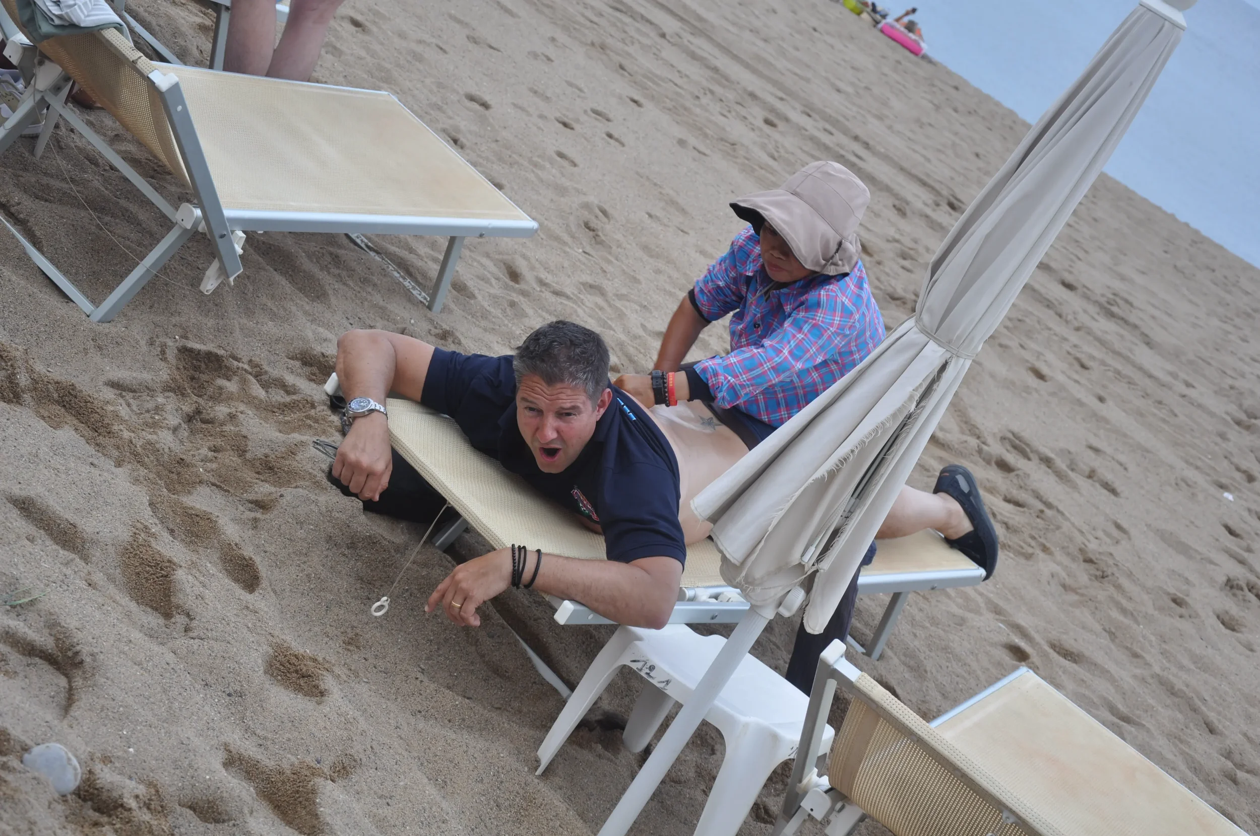 A man lying on a beach lounge chair with his head and upper body hanging over the edge, looking surprised. A person wearing a hat and plaid shirt is standing behind him, appearing to pull or push him. There are other lounge chairs and umbrellas on th