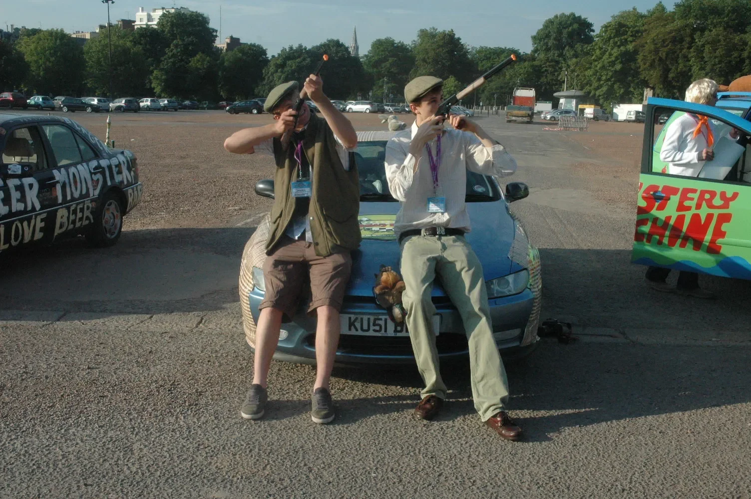 Two young men dressed in vintage-style clothing holding toy rifles, posing as if shooting, while leaning against the hood of a parked car in a parking lot. Other cars and a colorful vehicle with the words 'MYSTERY SHINE' are visible in the background