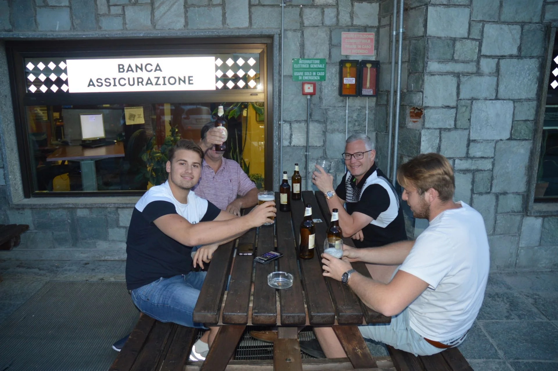 Group of five men sitting at a wooden table outside a building with a sign that says 'Banca Assicurazione', holding drinks and smiling. Inside the building, the window shows a workspace.