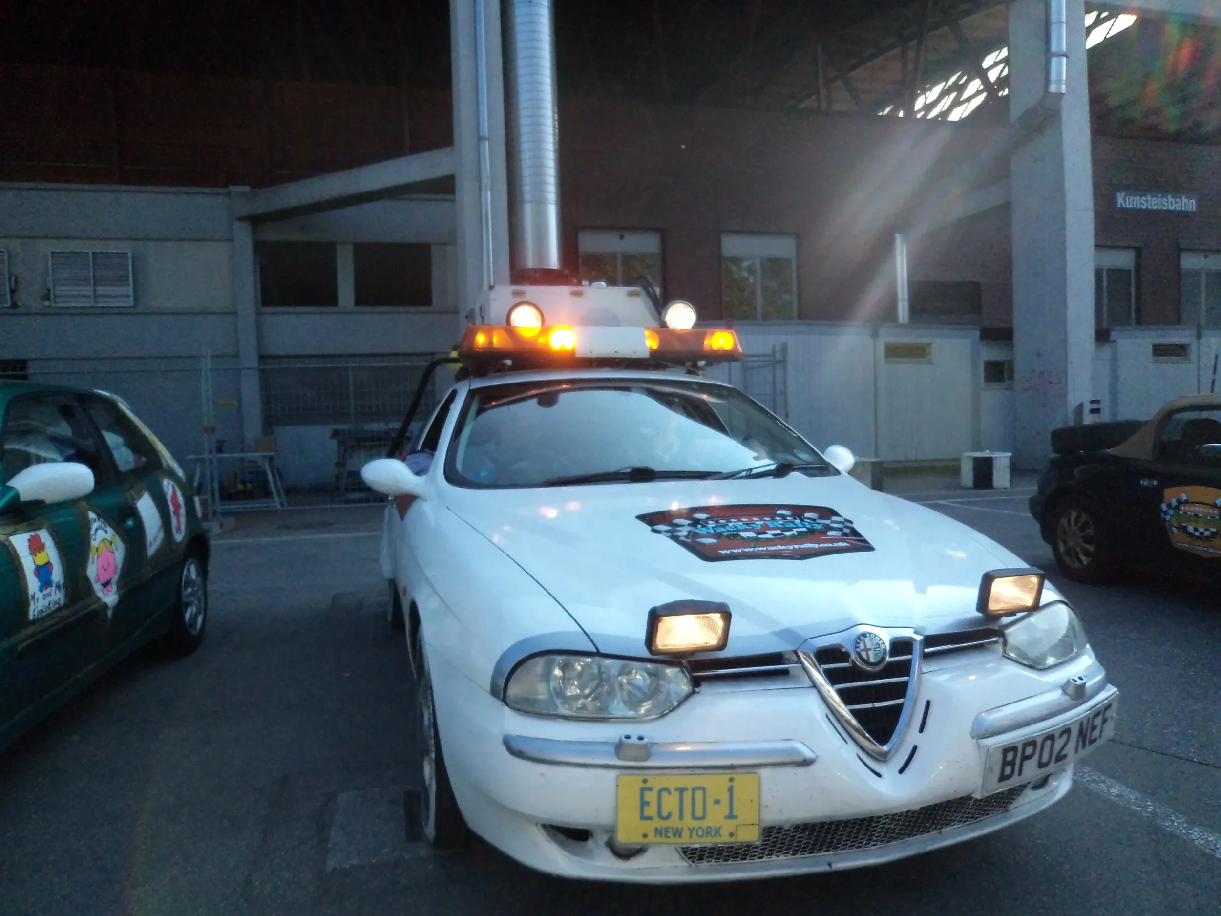 A white Alfa Romeo car with a light bar and a sign on the hood that reads 'Traffic Police' parked in a lot, with other cars around and a building in the background.