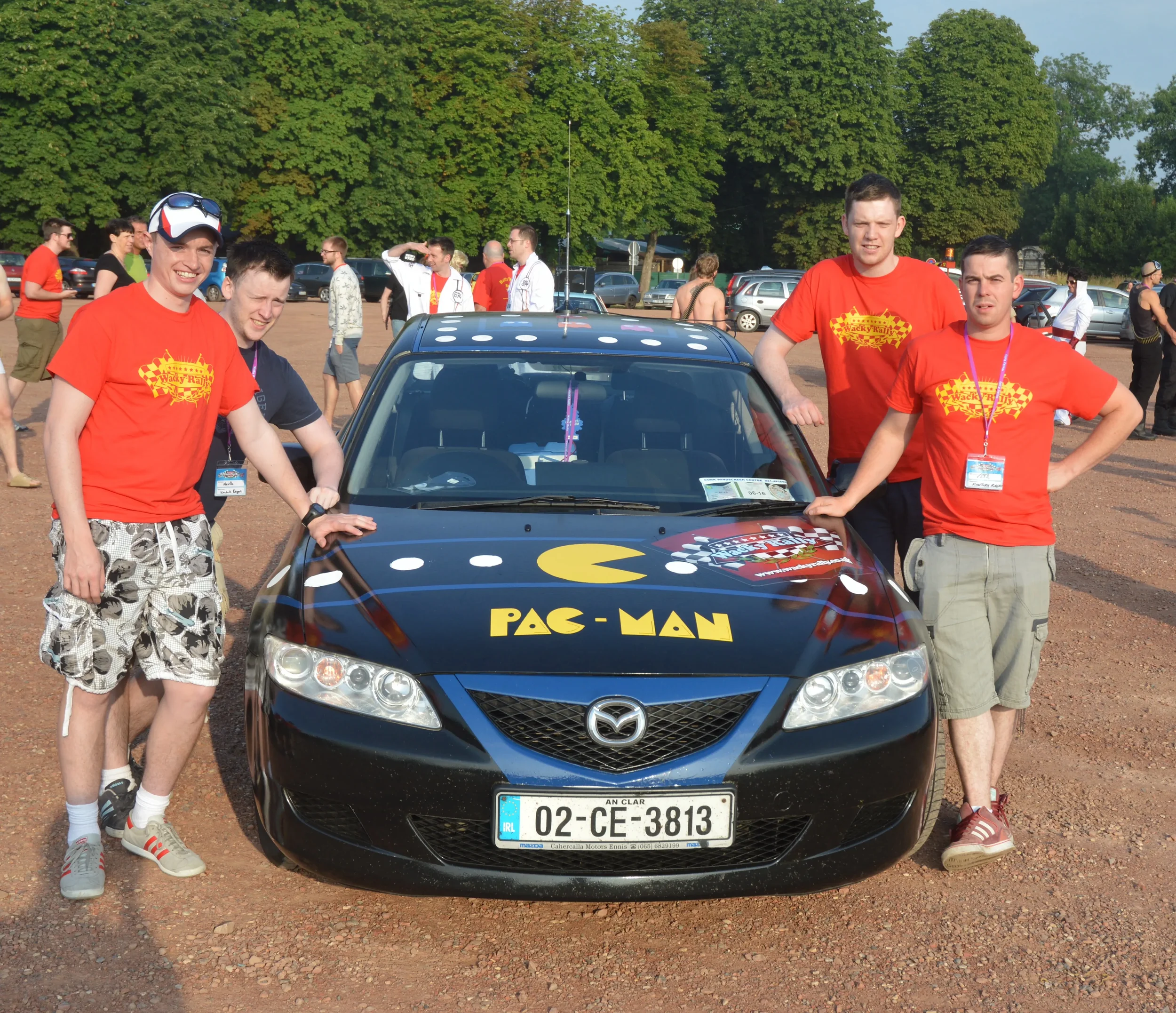 Group of four young men standing next to a black Mazda car decorated with Pac-Man graphics and characters from the classic video game, at an outdoor event.