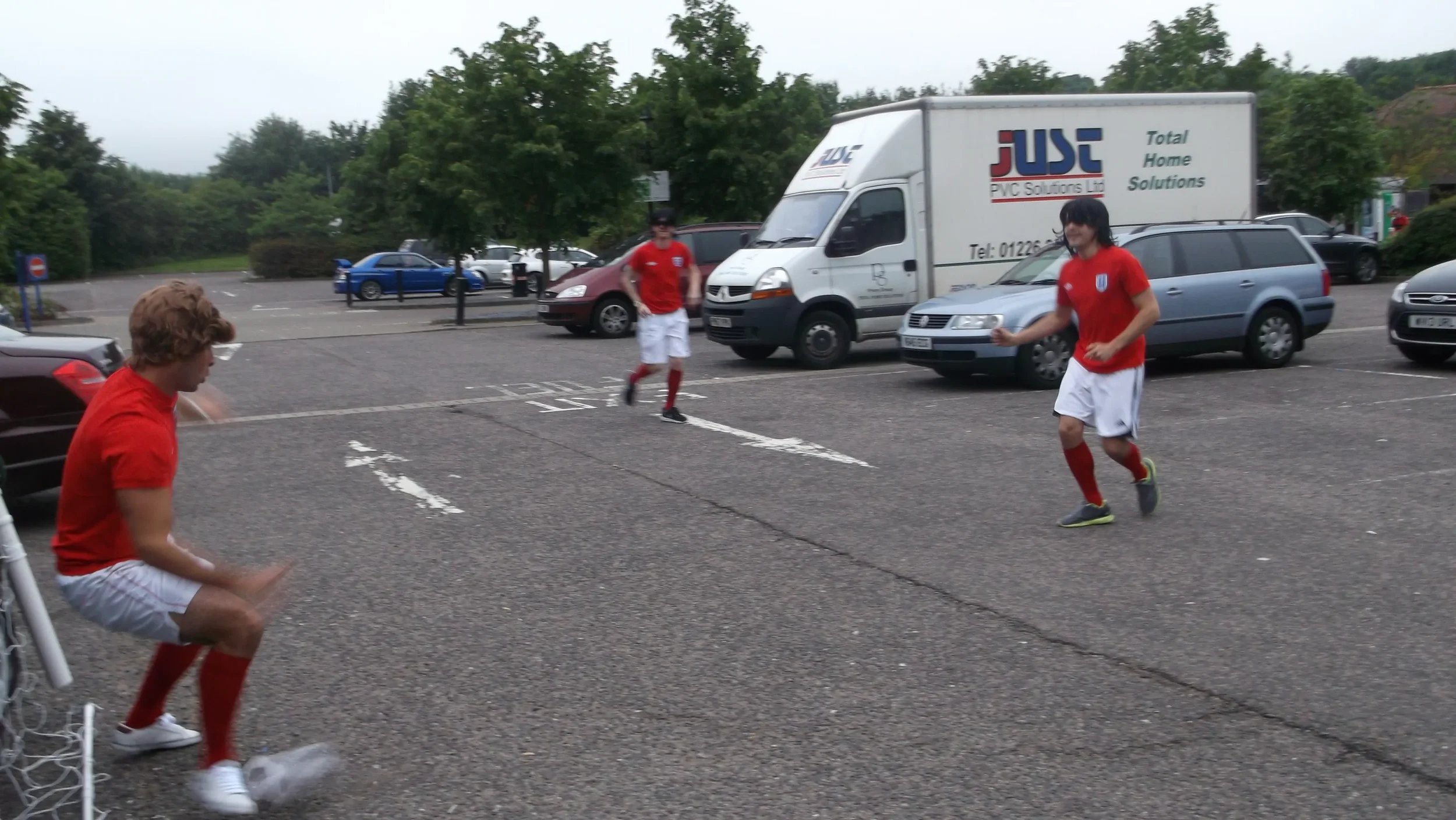 Three young men in red and white sports uniforms playing soccer in a parking lot.
