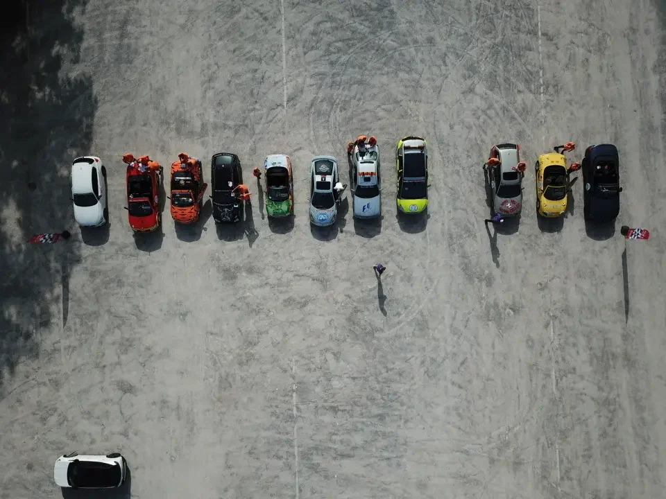 Aerial view of a row of 11 parked sports cars on a sandy area, with people standing around and walking near them. The cars are in various colors, and the area appears to be a racing or car event setting.