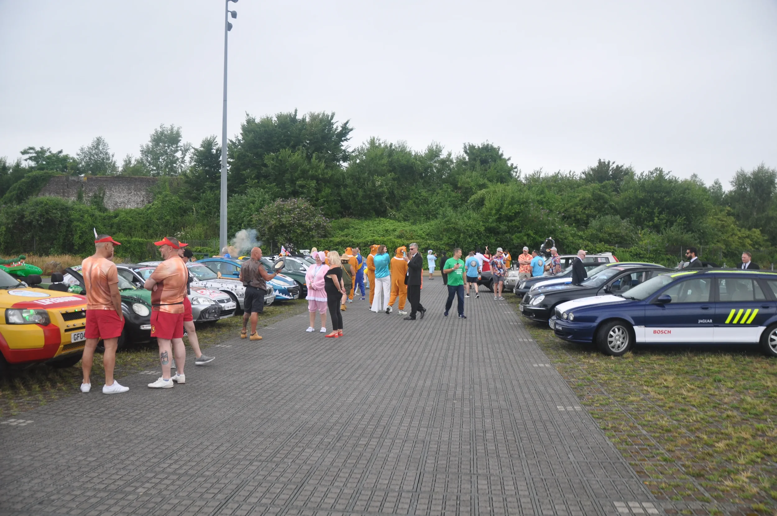 People dressed in various casual and costume outfits gather in a parking lot, surrounded by vintage and modern cars, with greenery in the background.
