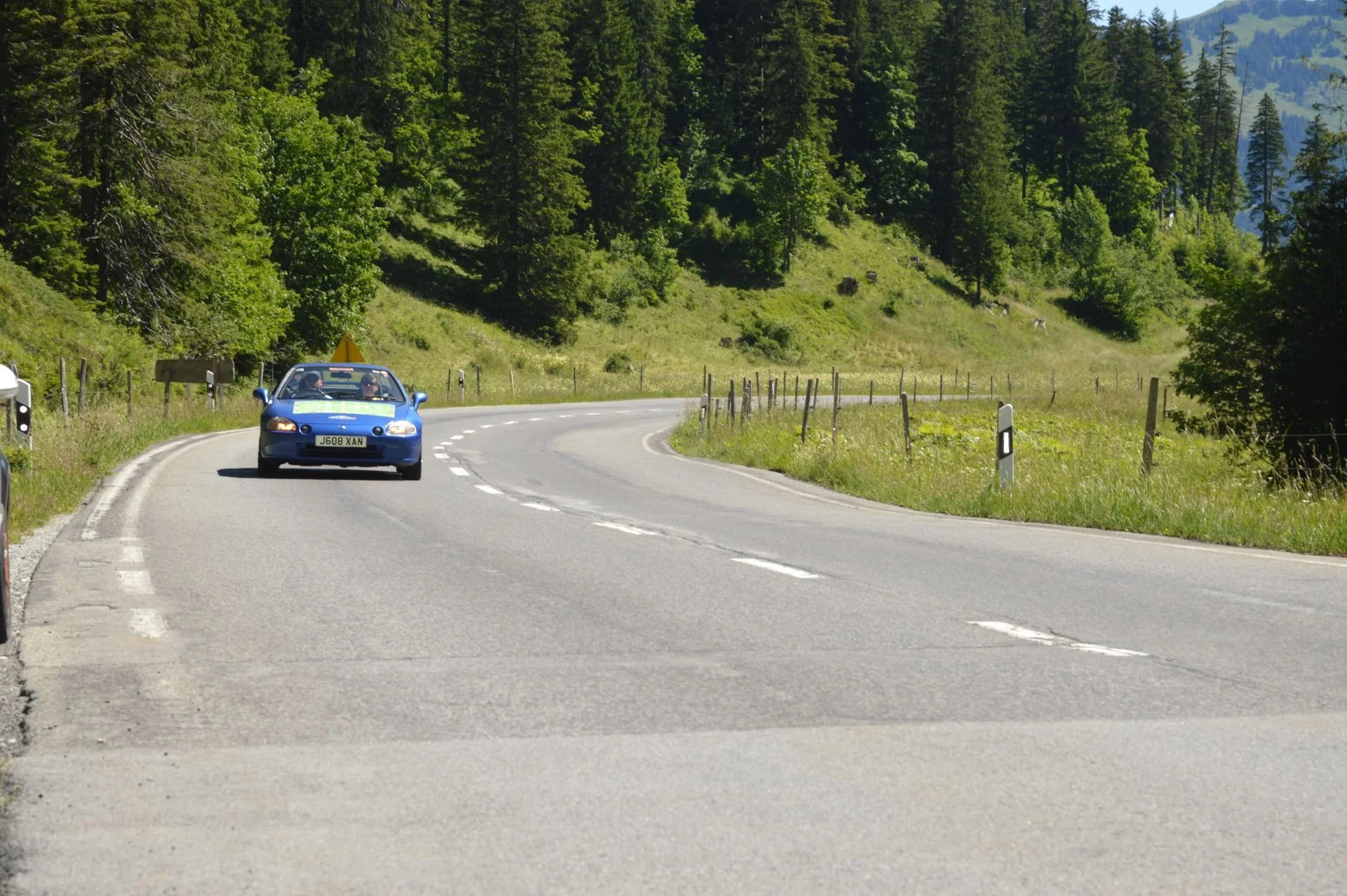 A winding mountain road with a blue car driving around a bend, in a scenic landscape with trees and hills in the background.