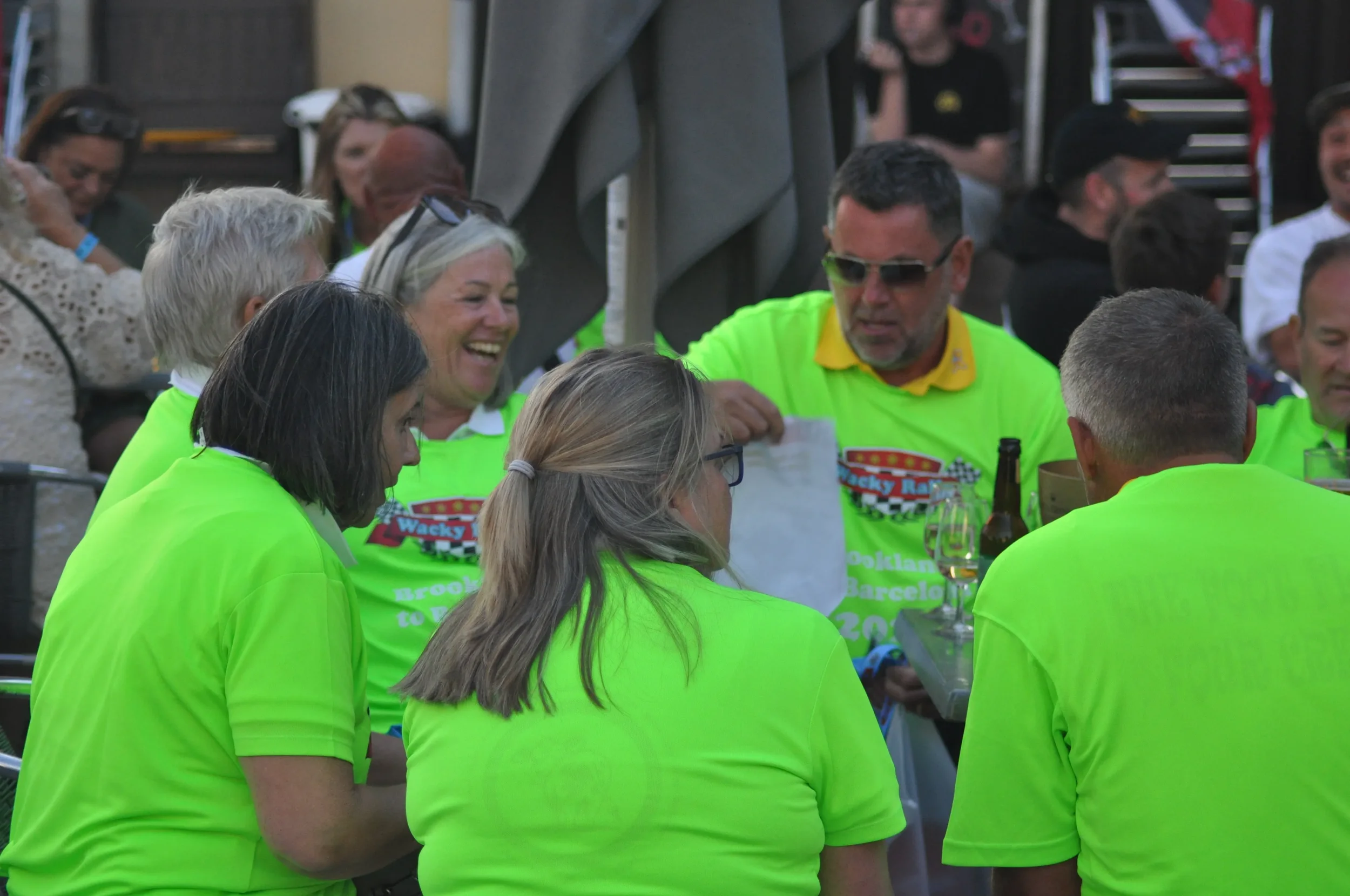 Group of people sitting together at a table, some of them laughing and smiling. They are wearing bright yellow-green shirts with a racing logo. The table has drinks, including a glass of beer and a glass of champagne.