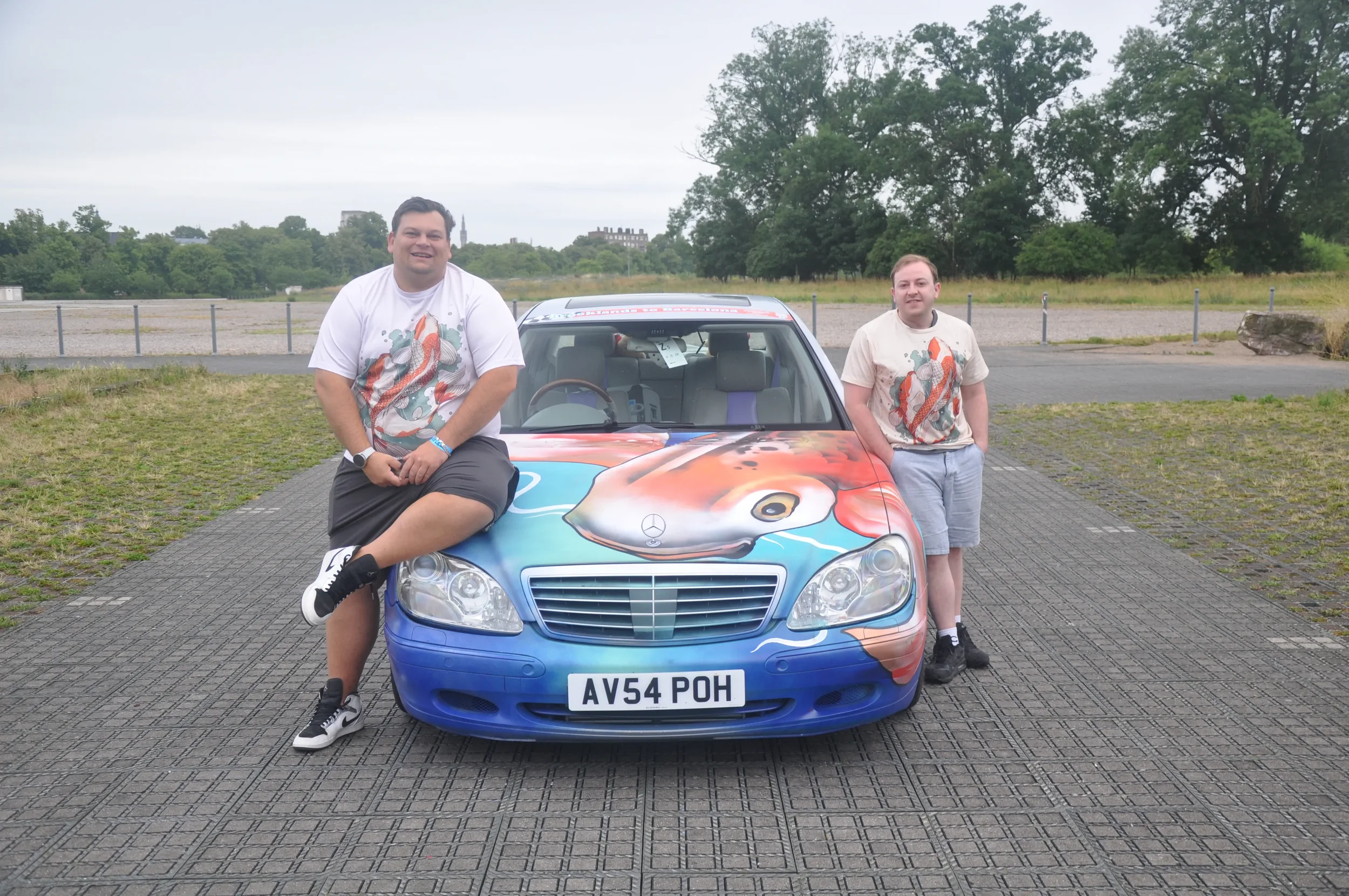 Two men with tattoos wearing matching T-shirts with a koi fish design, standing beside a Mercedes-Benz car with a large koi fish decal on the hood. The setting is an outdoor parking lot with trees and open space in the background.