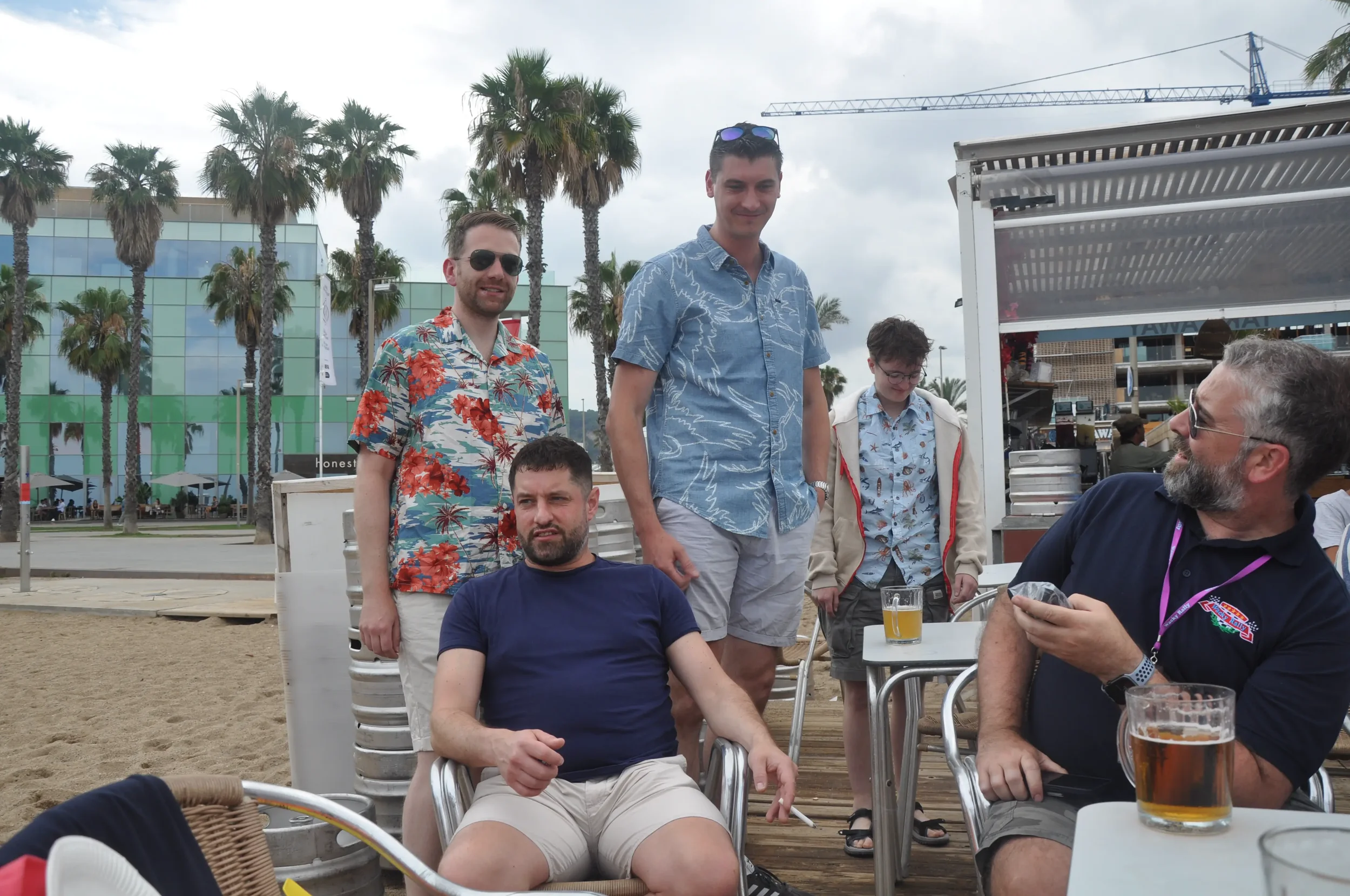 Group of people at a beachside outdoor bar, some sitting, others standing, with palm trees and a modern glass building in the background. Two men are seated holding drinks, and the others are standing behind them, chatting and smiling.