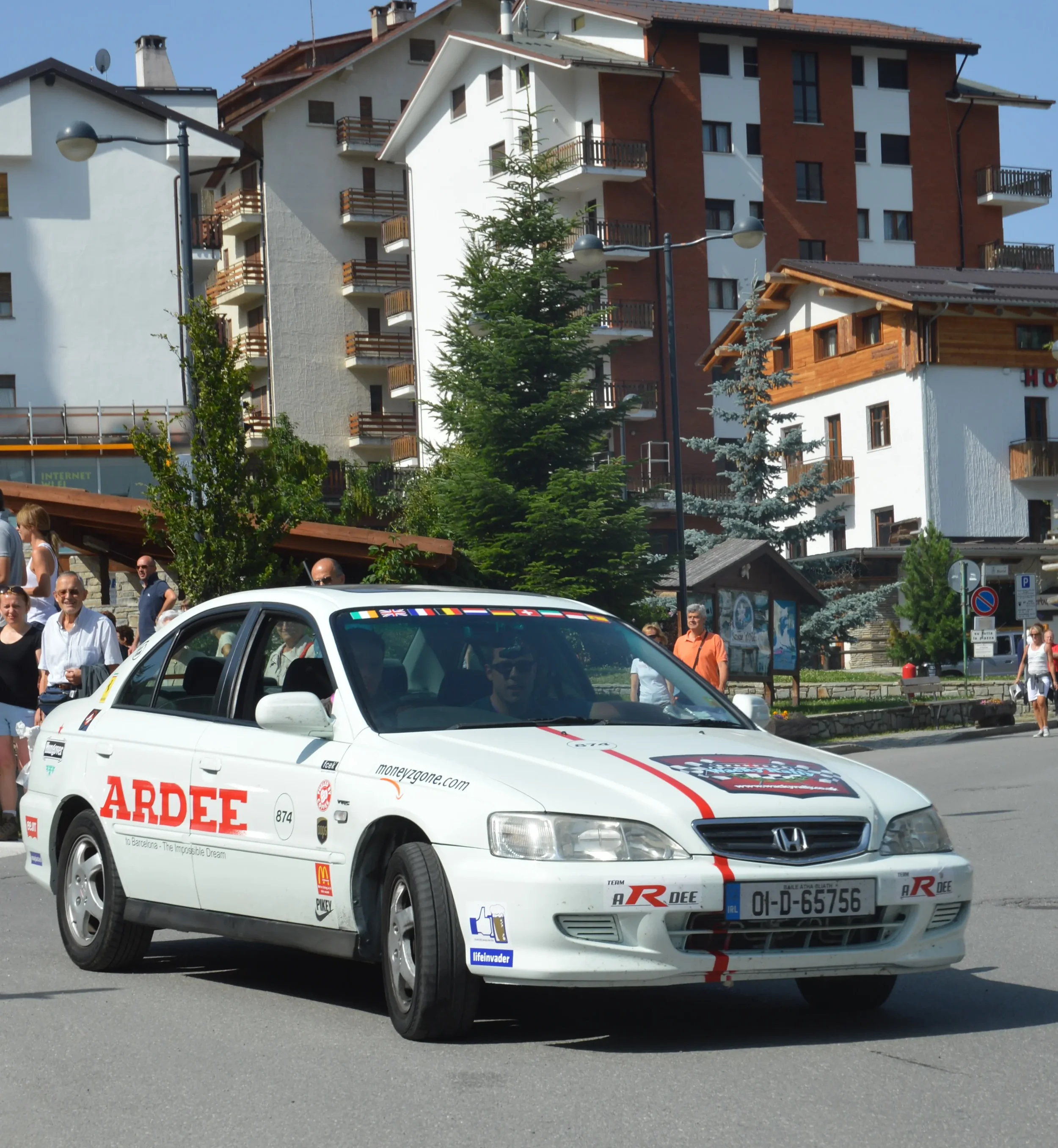 A white Honda sedan with various stickers and decals, including 'ARDEE', is driving on a street in a town with trees and buildings in the background, and several people are walking on the sidewalk.
