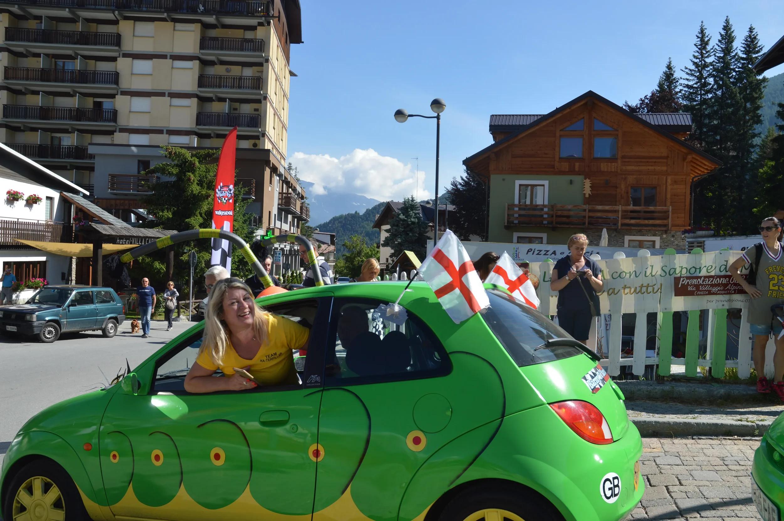 A smiling woman with blonde hair leaning out of a bright green cartoon-style car decorated with snake patterns, during a parade in a mountain town, with several spectators and buildings in the background.