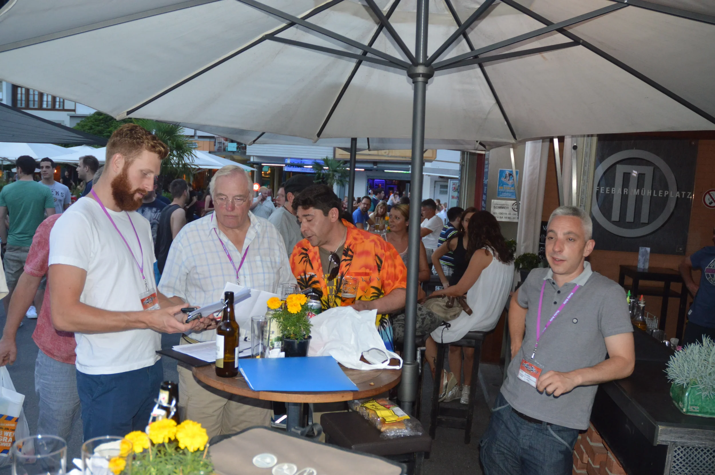 A group of men gathered around a table under a large patio umbrella at an outdoor event. They are engaged in conversation, some holding papers and drinks, with a crowd of people in the background.