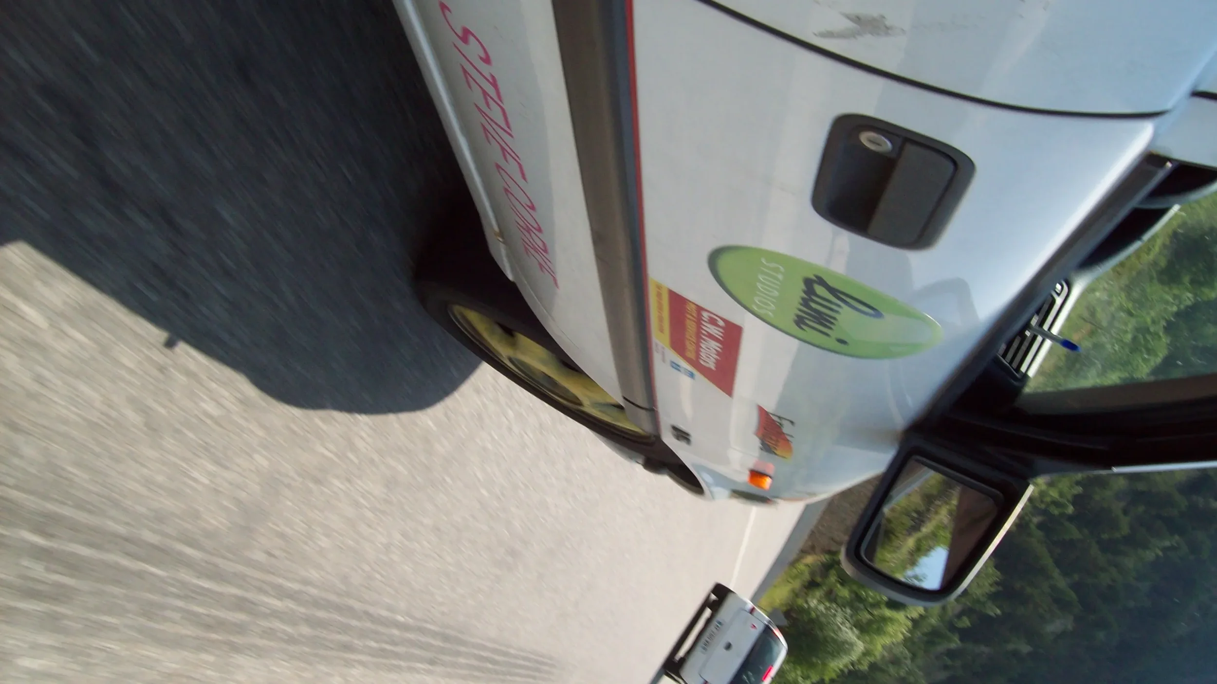 A white vehicle with a green and yellow logo on the side, driving on a highway with trees in the background.