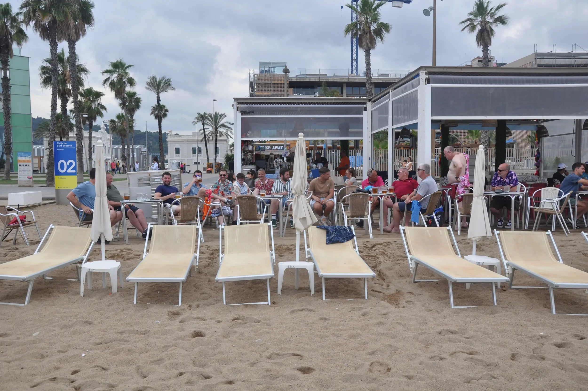 Group of people sitting at tables and on chairs on a beach with sand, palm trees, and cloudy sky.