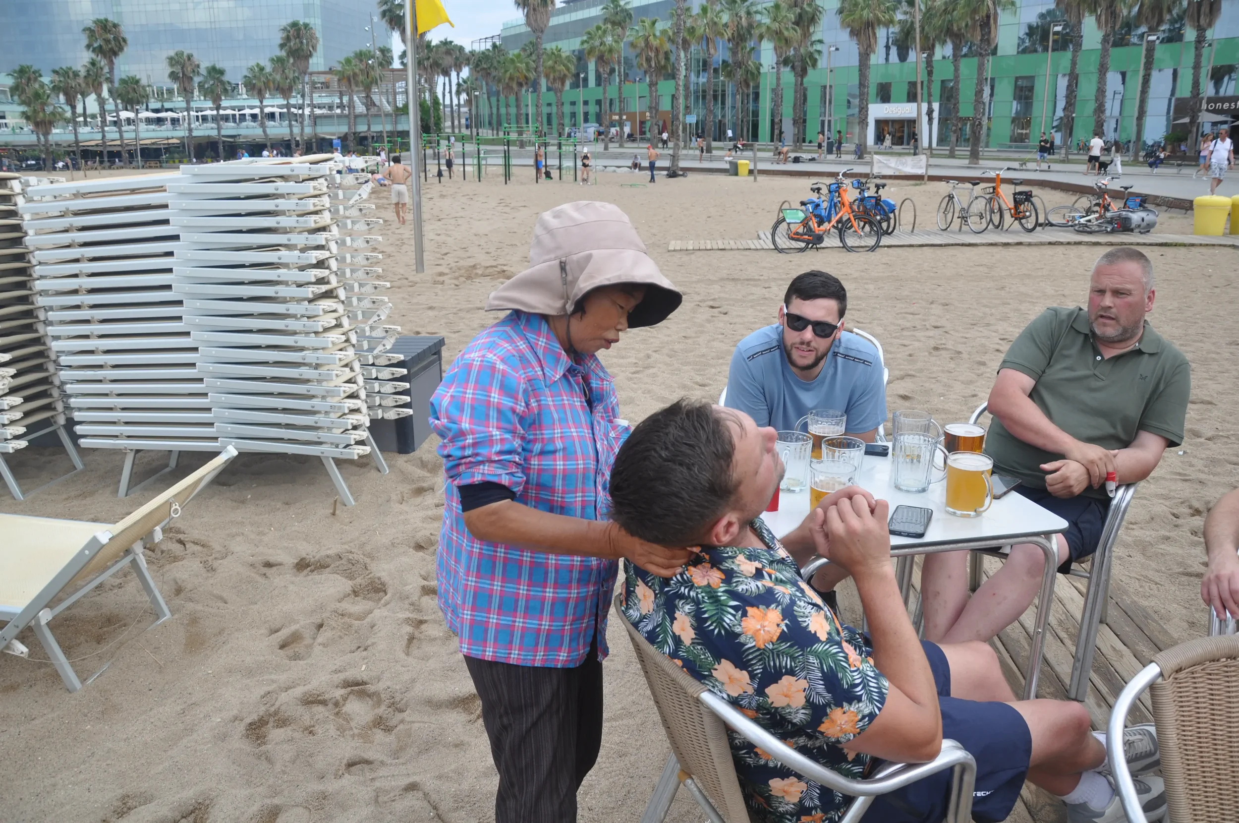 People gathered at a table on a sandy beach, with one woman giving a neck massage to a man. There are four glasses of beer on the table, and in the background, there are multiple bicycles, palm trees, and a modern glass building.