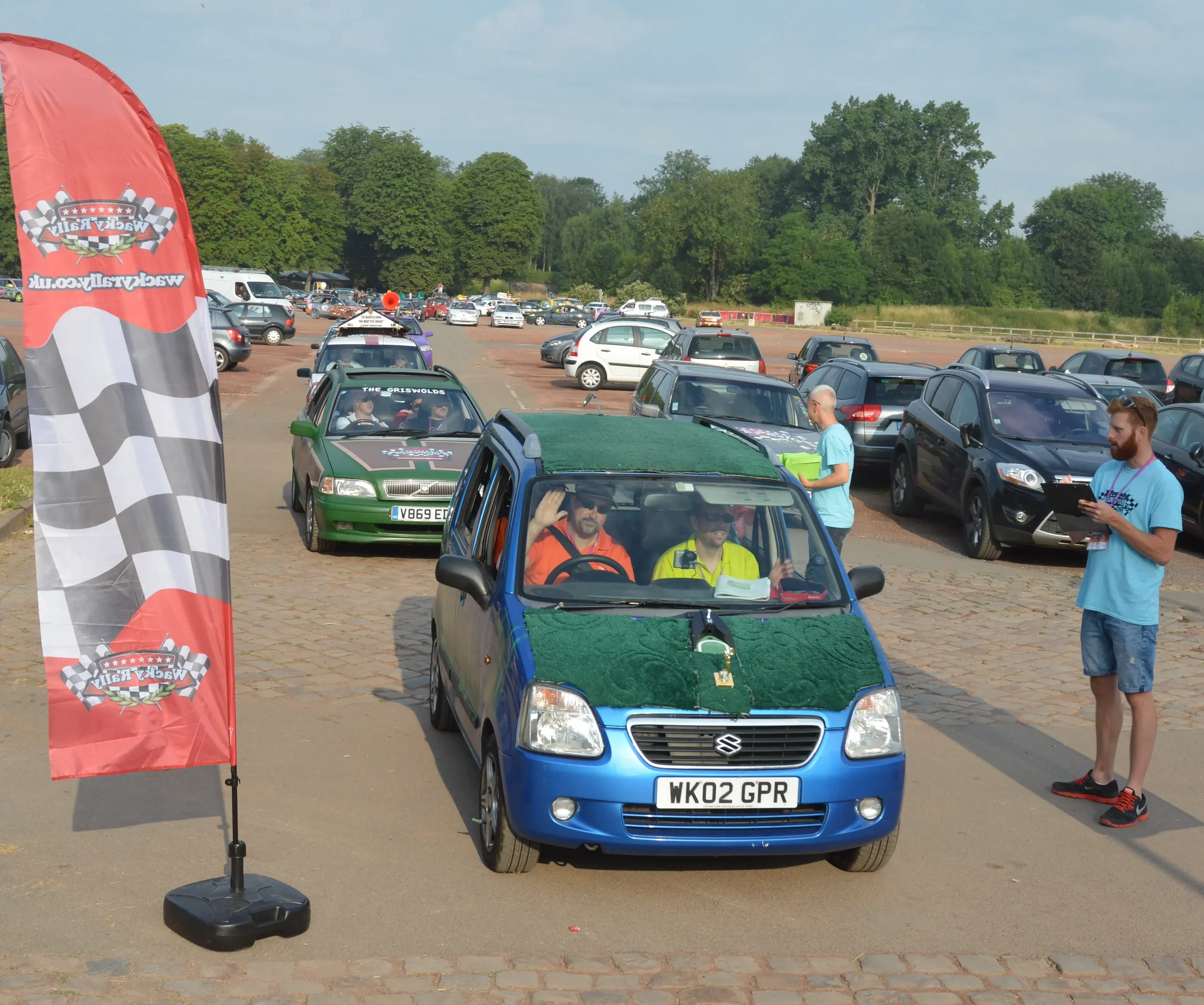 A blue Suzuki car decorated with green mats on the hood and windshield, parked at a car event with people around, a red flag with a checkered flag pattern and the event logo, and other cars in the background.
