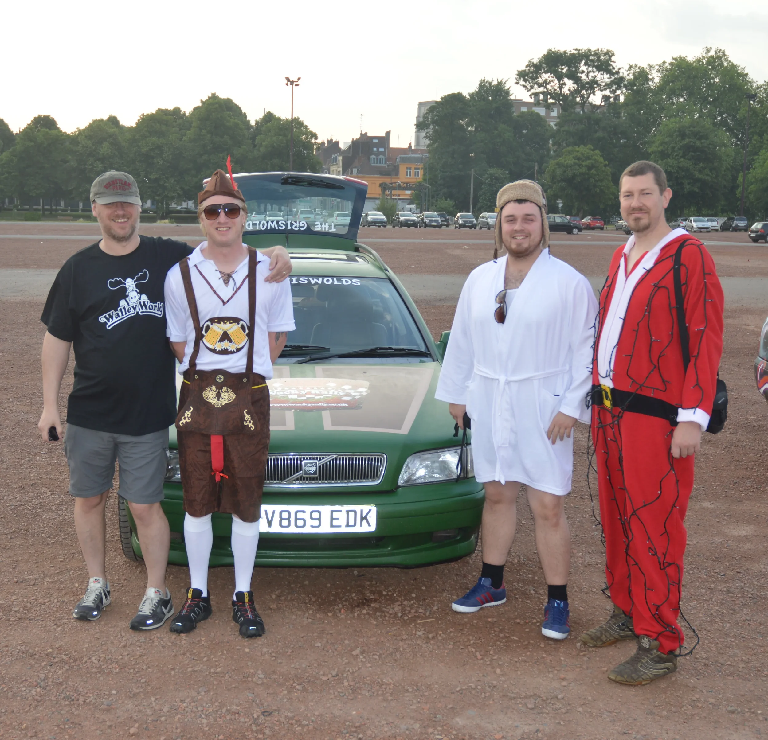 Four men dressed in costumes standing in front of a green car on an outdoor dirt area with trees and buildings in the background.