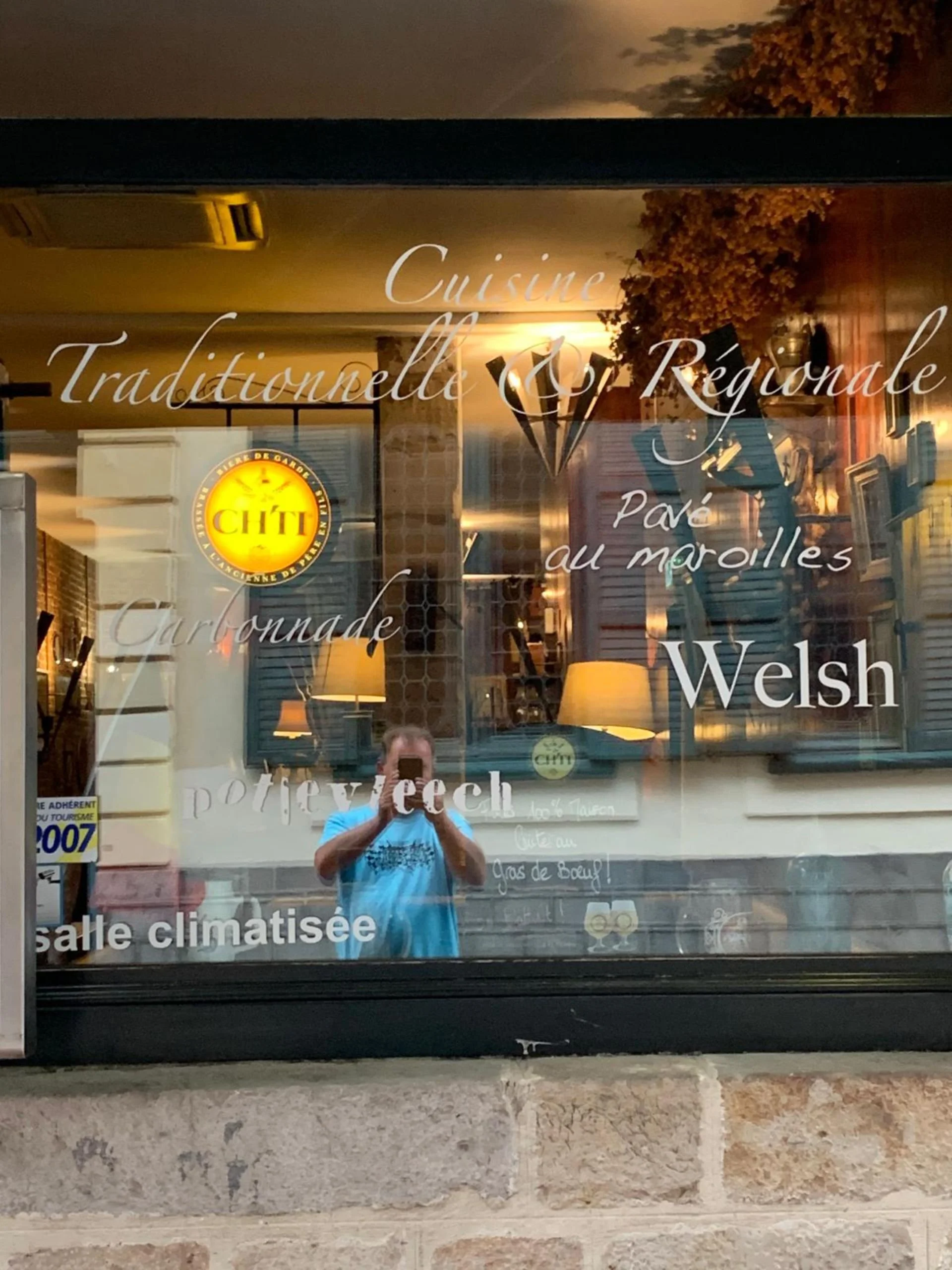 Reflection of a person taking a photo in a glass window of a restaurant exterior, with words in French listing cuisine types, a logo, and interior lighting visible.